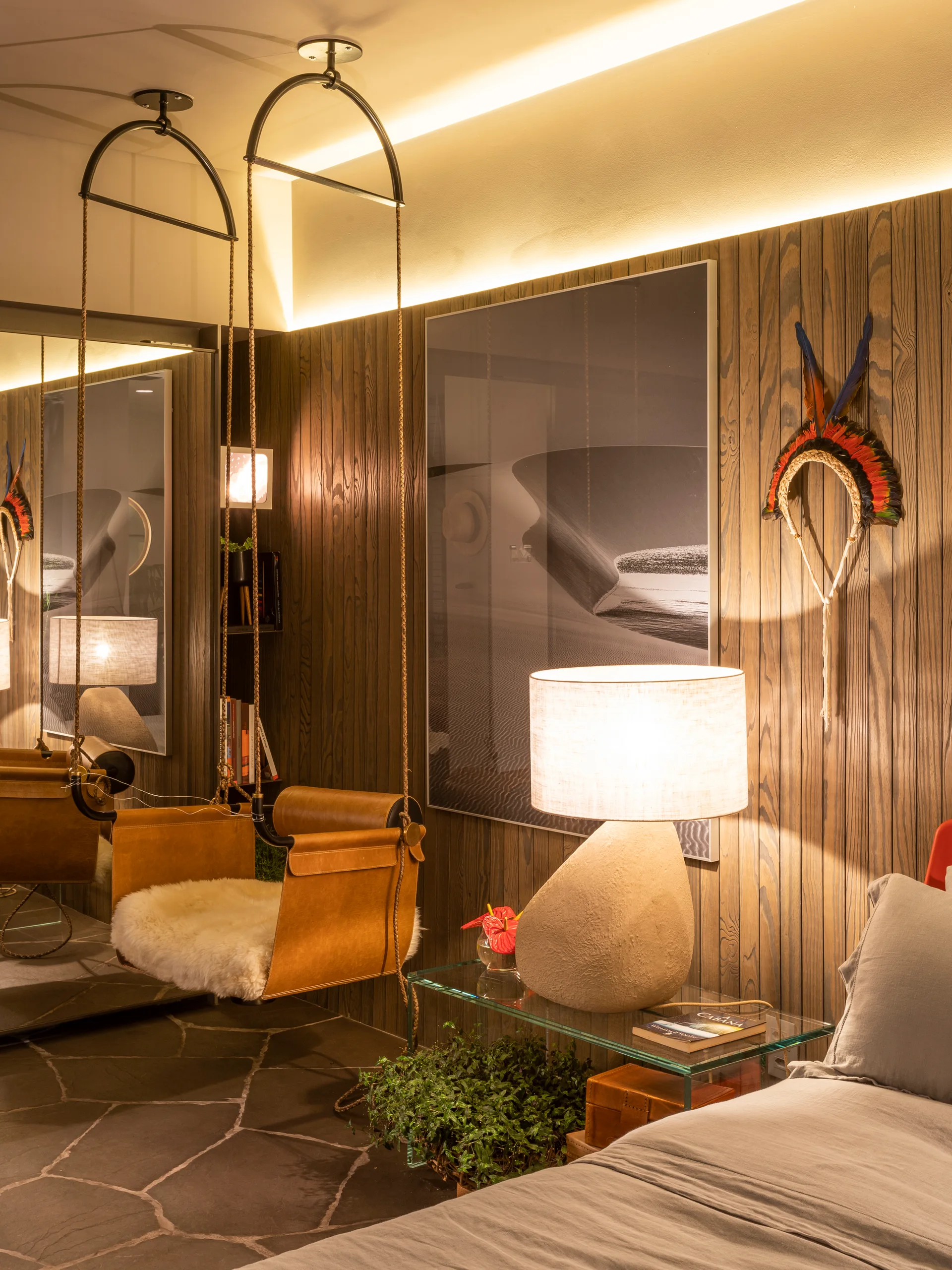 An organic modern bedroom with dark wood paneling, a suspended leather swing chair, and a sculptural table lamp on a glass nightstand.