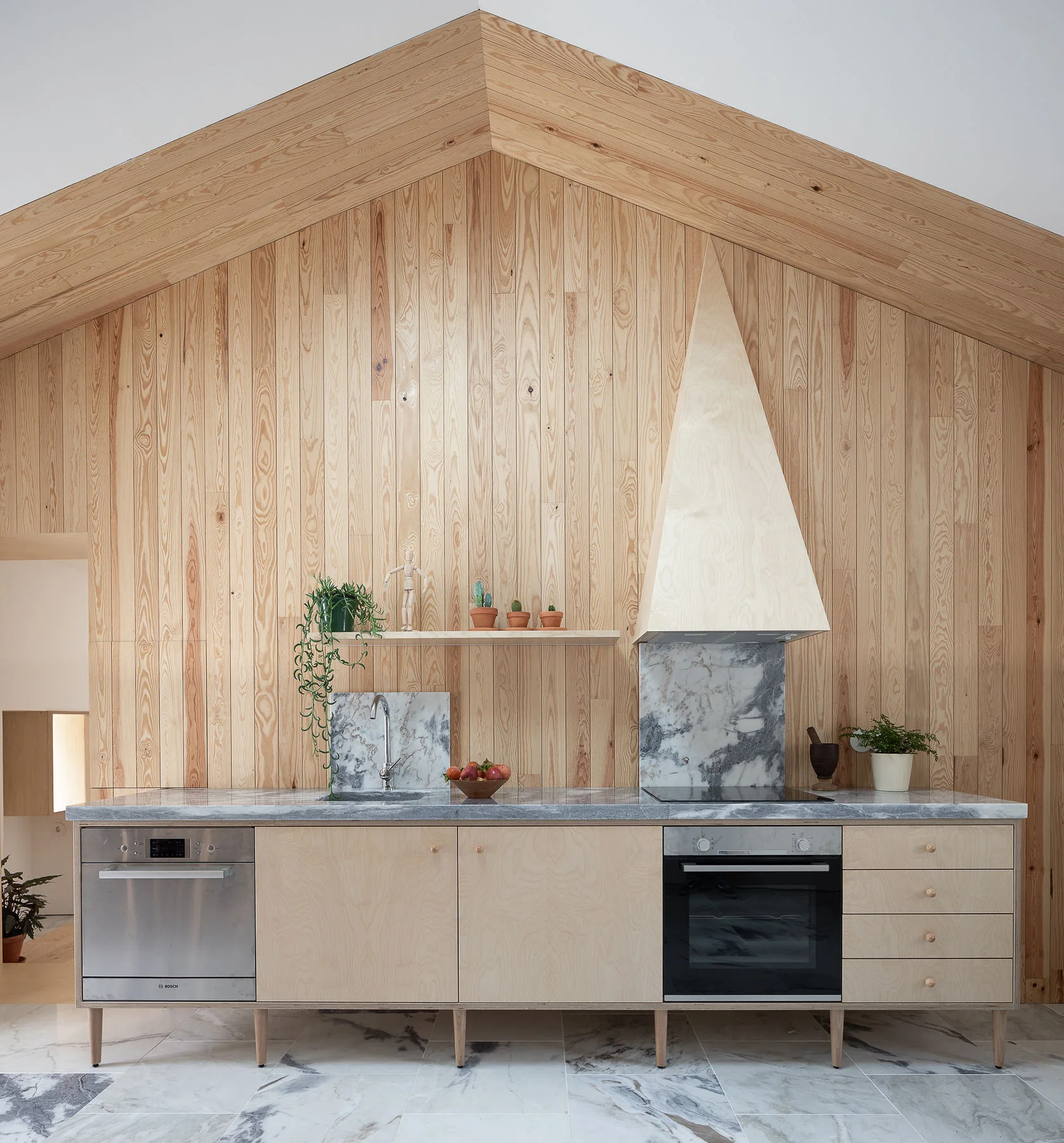A minimalist kitchen features vertical light wood paneling, a vaulted ceiling, light wood cabinets on legs, and grey veined marble countertops.