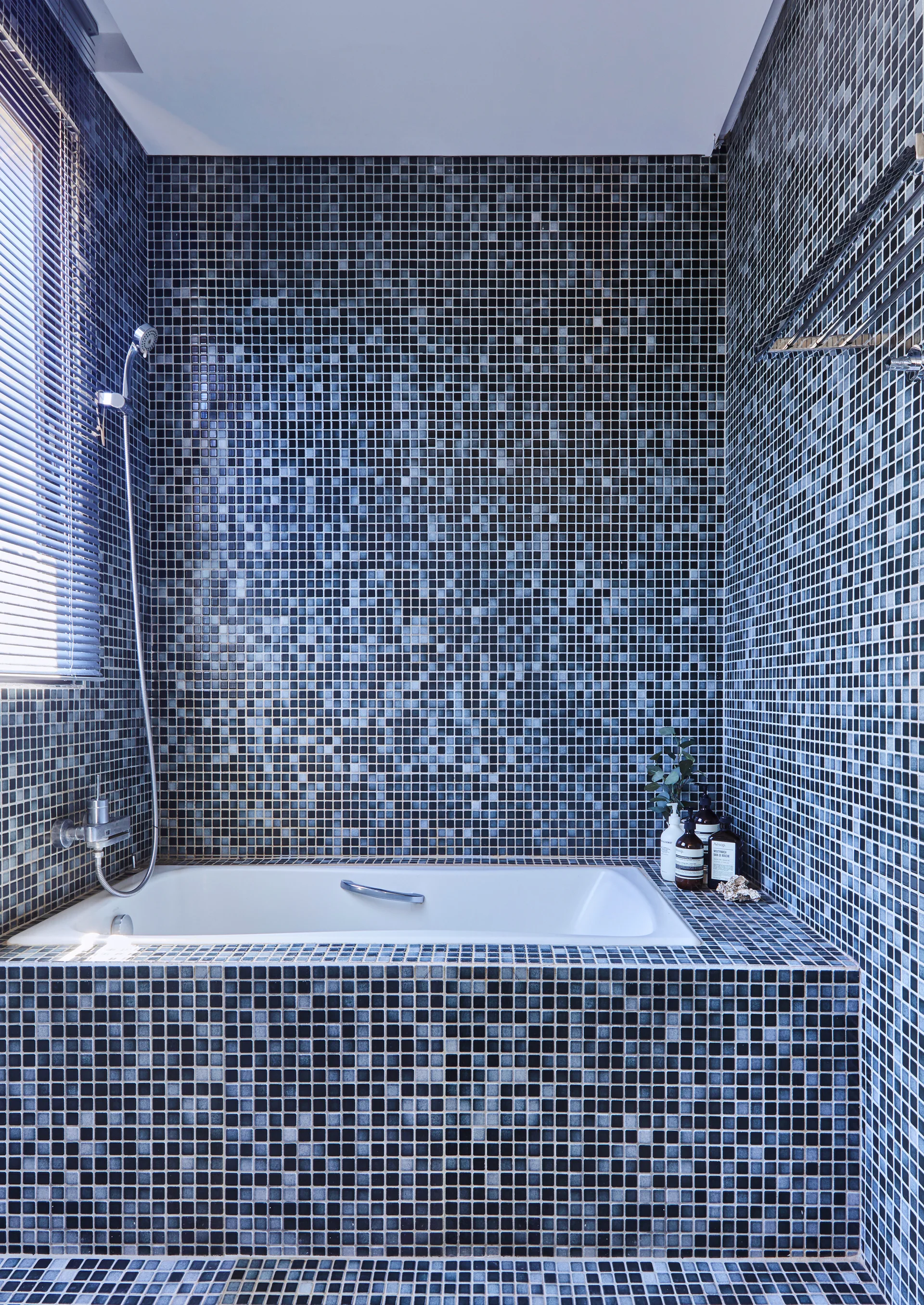 A white built-in bathtub surrounded by walls and a deck covered in dark blue and black mosaic tiles next to a window with blinds.