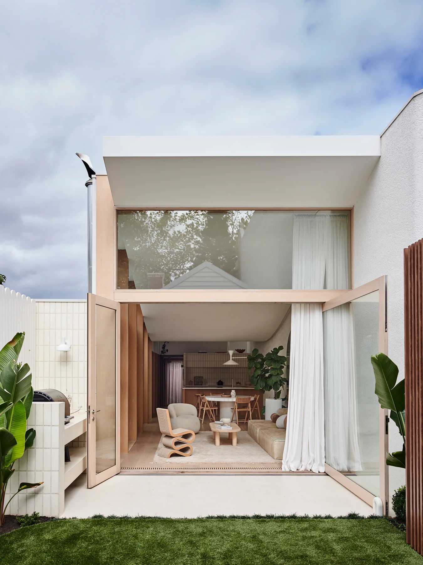 Large wood-framed glass doors open from a concrete patio with a built-in grill into a neutral living room with a sculptural chair.