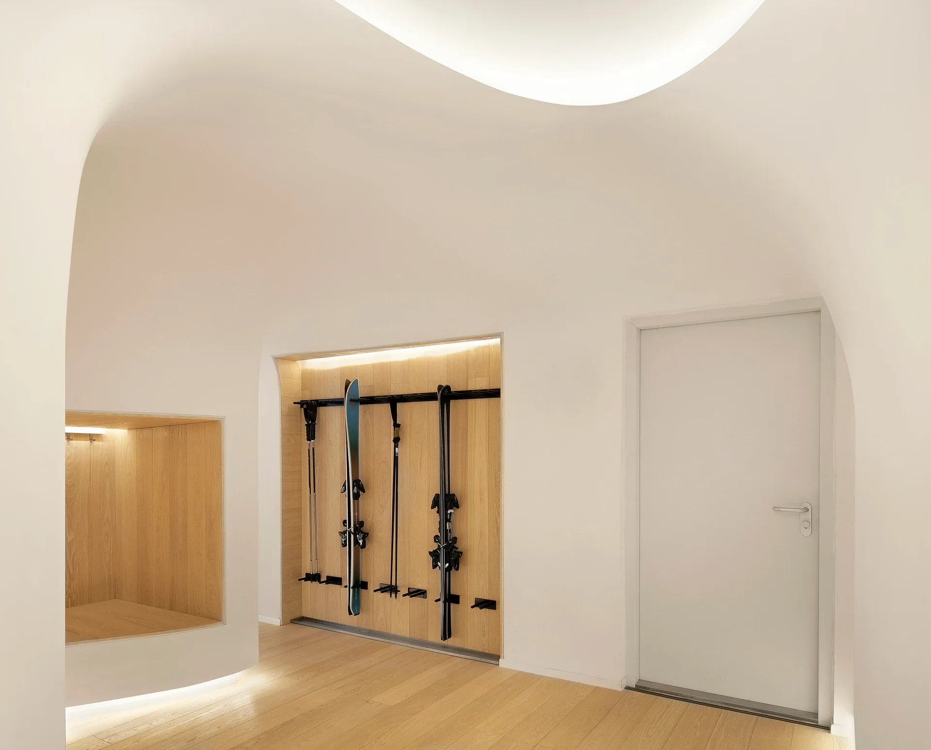 A minimalist white mudroom featuring organic curved walls, light wood flooring, a built-in wooden ski storage niche, and cove lighting.