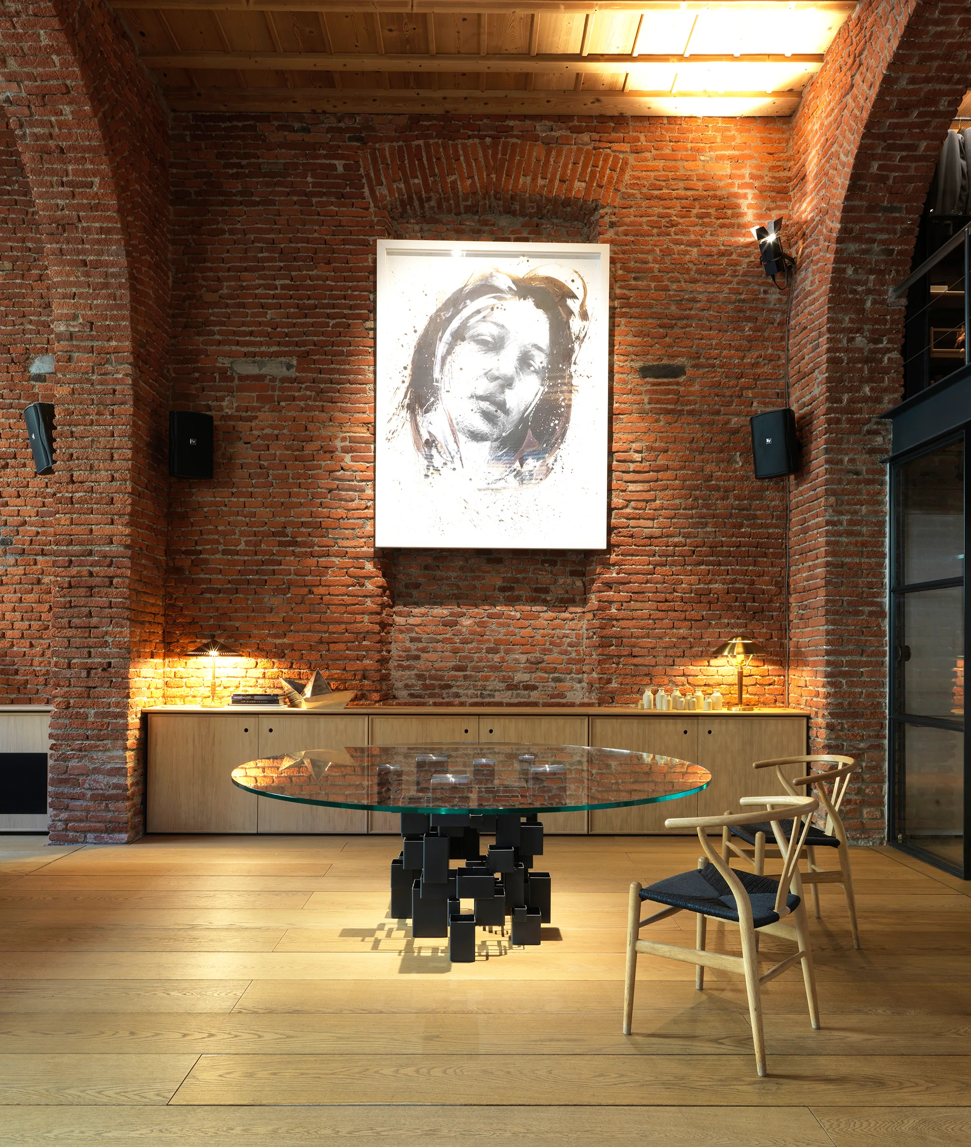 A round glass dining table with a sculptural base stands on wood floors in front of a low cabinet and an exposed brick wall.