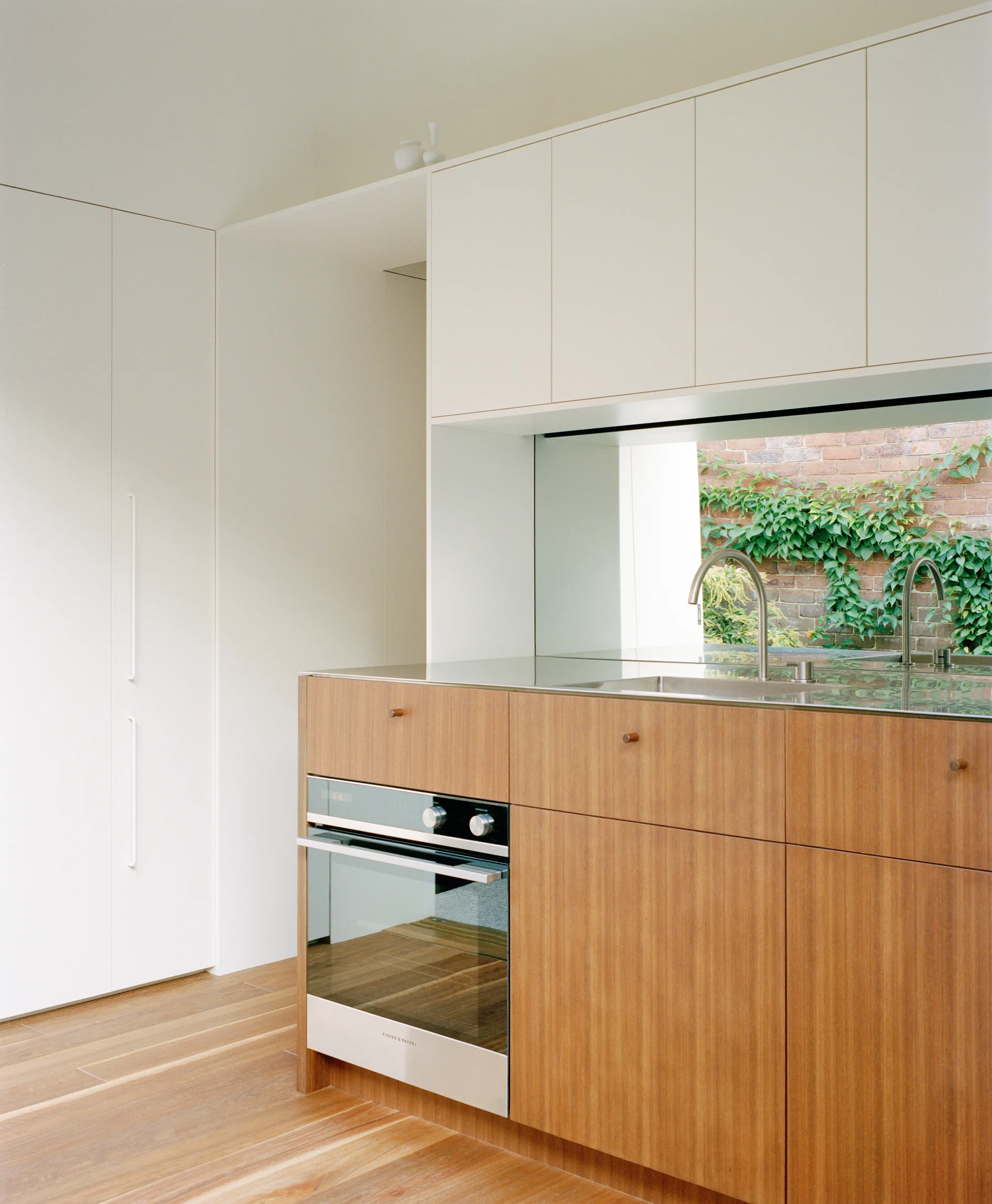 A minimalist kitchen with white upper cabinets, wood-grained lower cabinets, and a mirrored backsplash reflecting an outdoor brick wall with ivy.