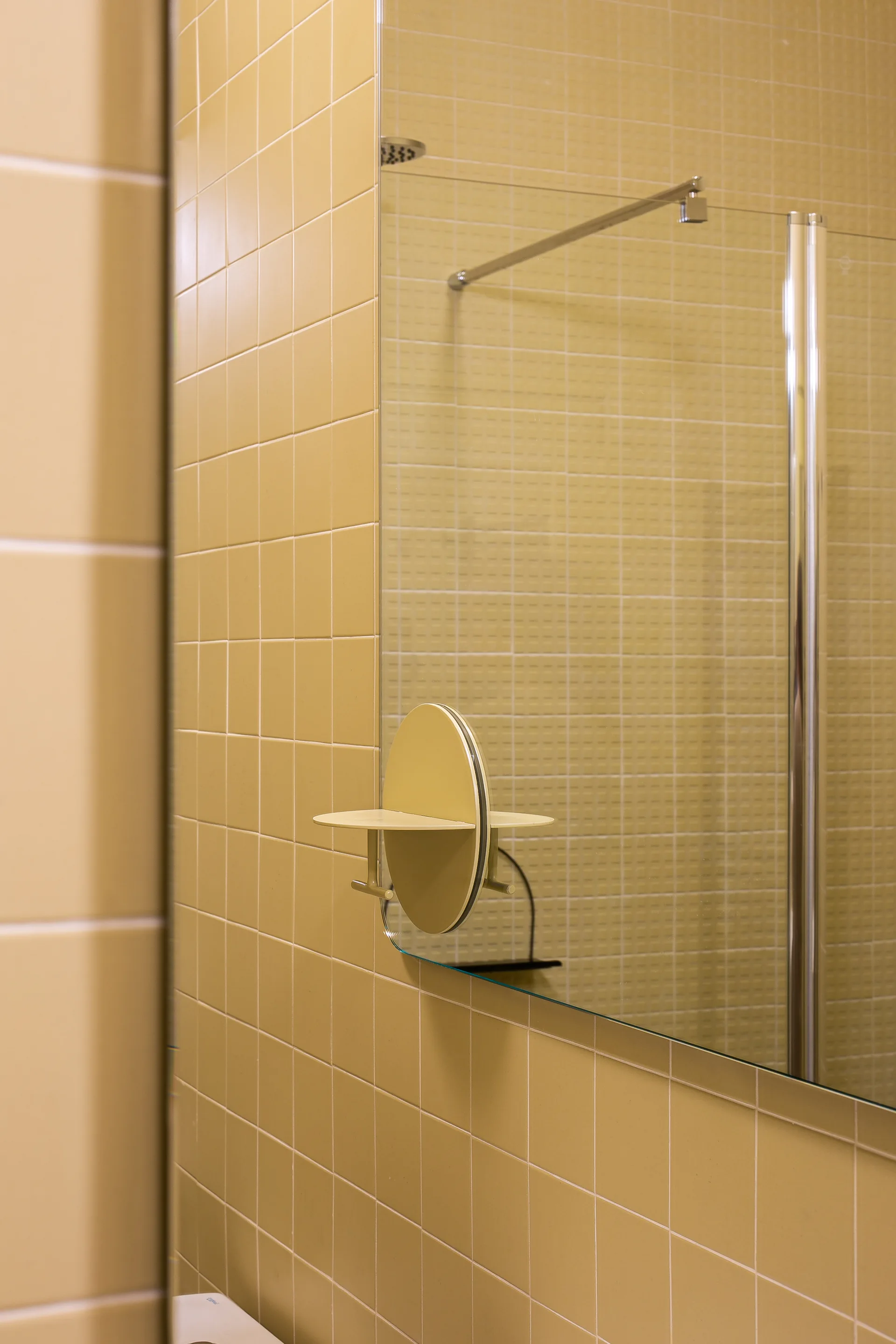 A bathroom wall covered in yellow square tiles features a frameless mirror with an integrated yellow circular shelf and a glass shower panel reflection.