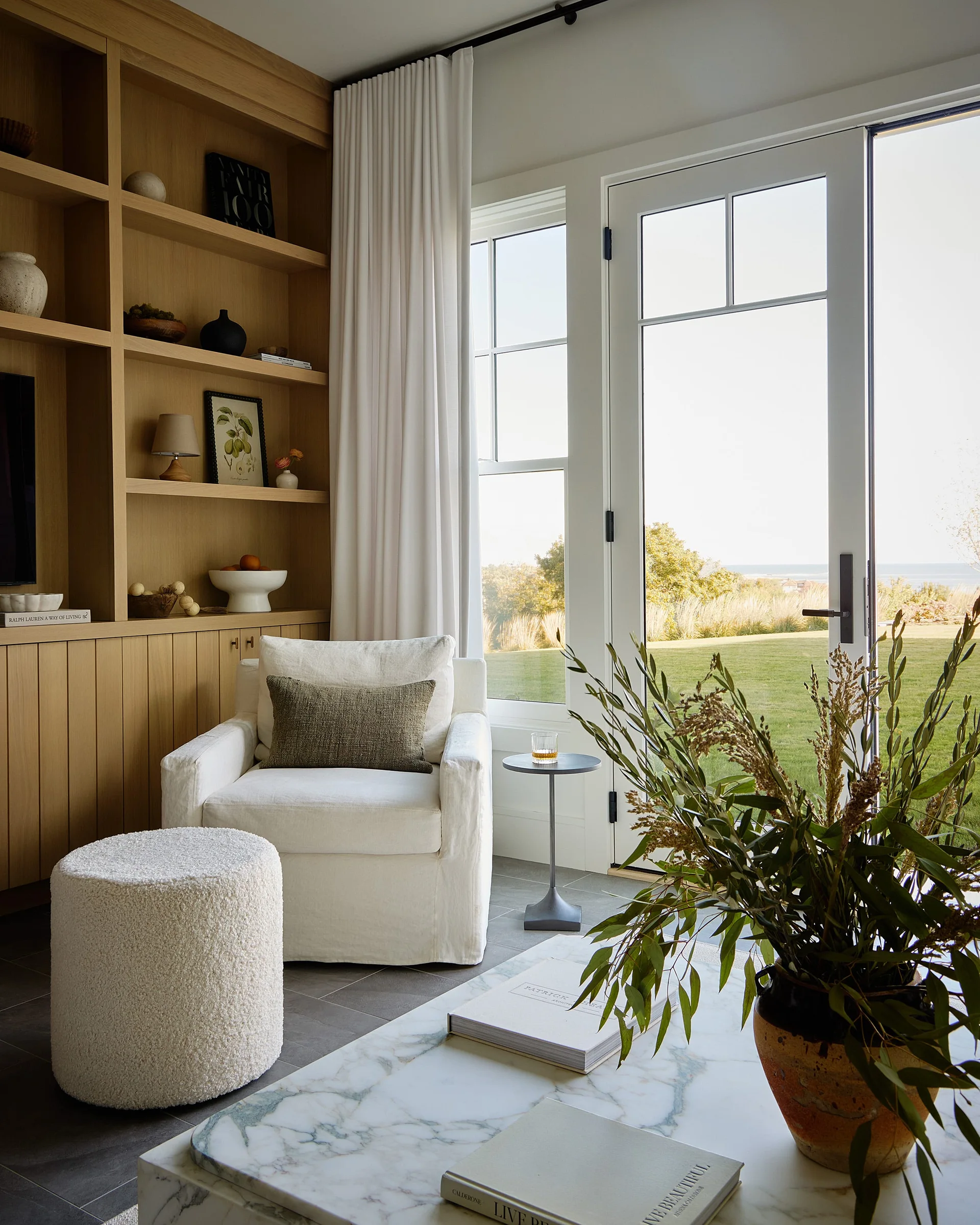 A white slipcovered armchair sits next to light wood built-in bookshelves and French doors that open to a green lawn.