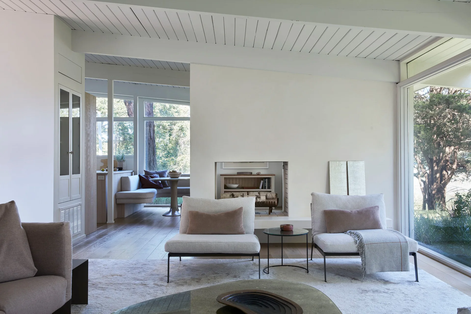 A minimalist living room features two low white armchairs, a central recessed fireplace, and a large window looking out onto trees.