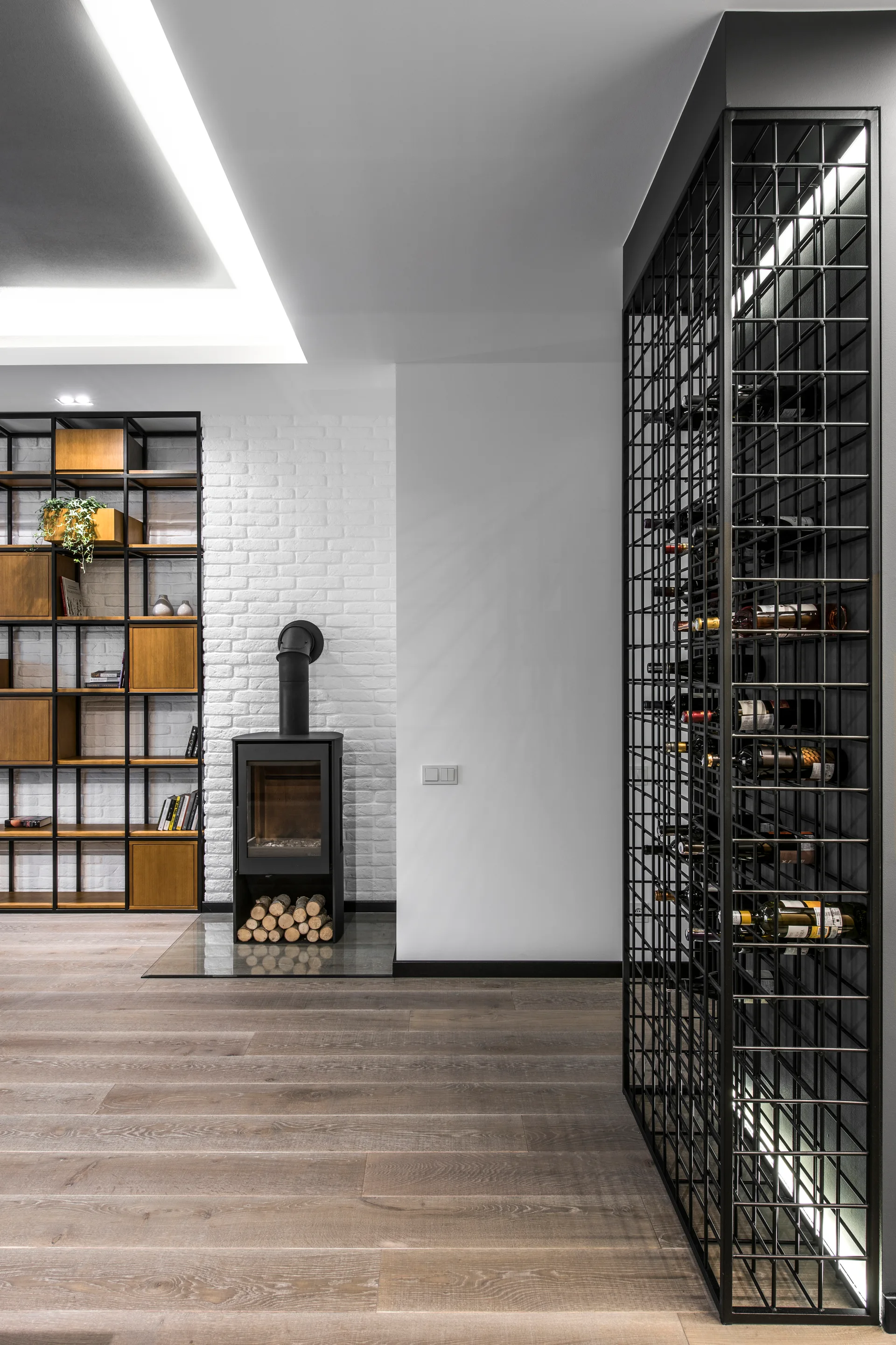 A modern interior space featuring a tall black metal grid wine rack, a freestanding fireplace against a white brick wall, and wood floors.