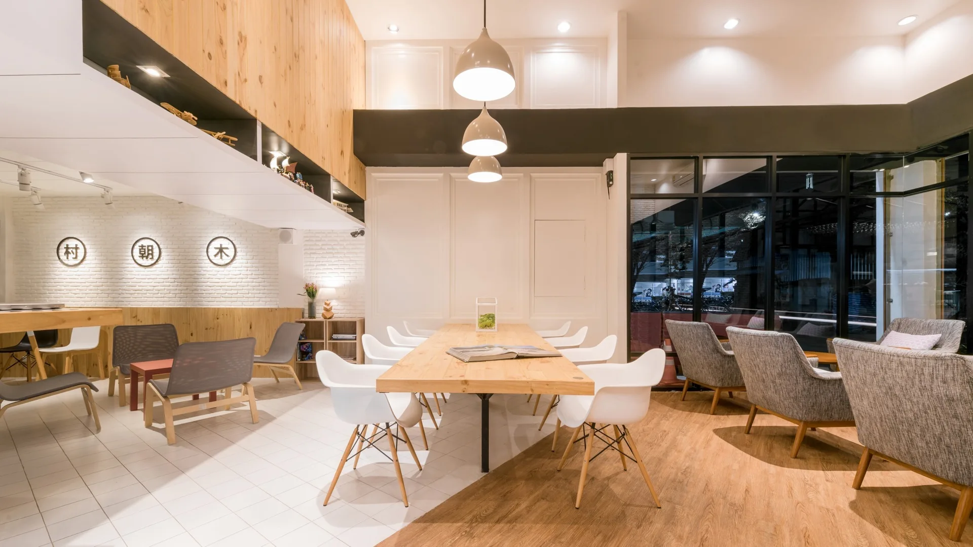 A spacious dining area features a long wooden table with white chairs on mixed tile and wood flooring, next to gray armchairs and large windows.