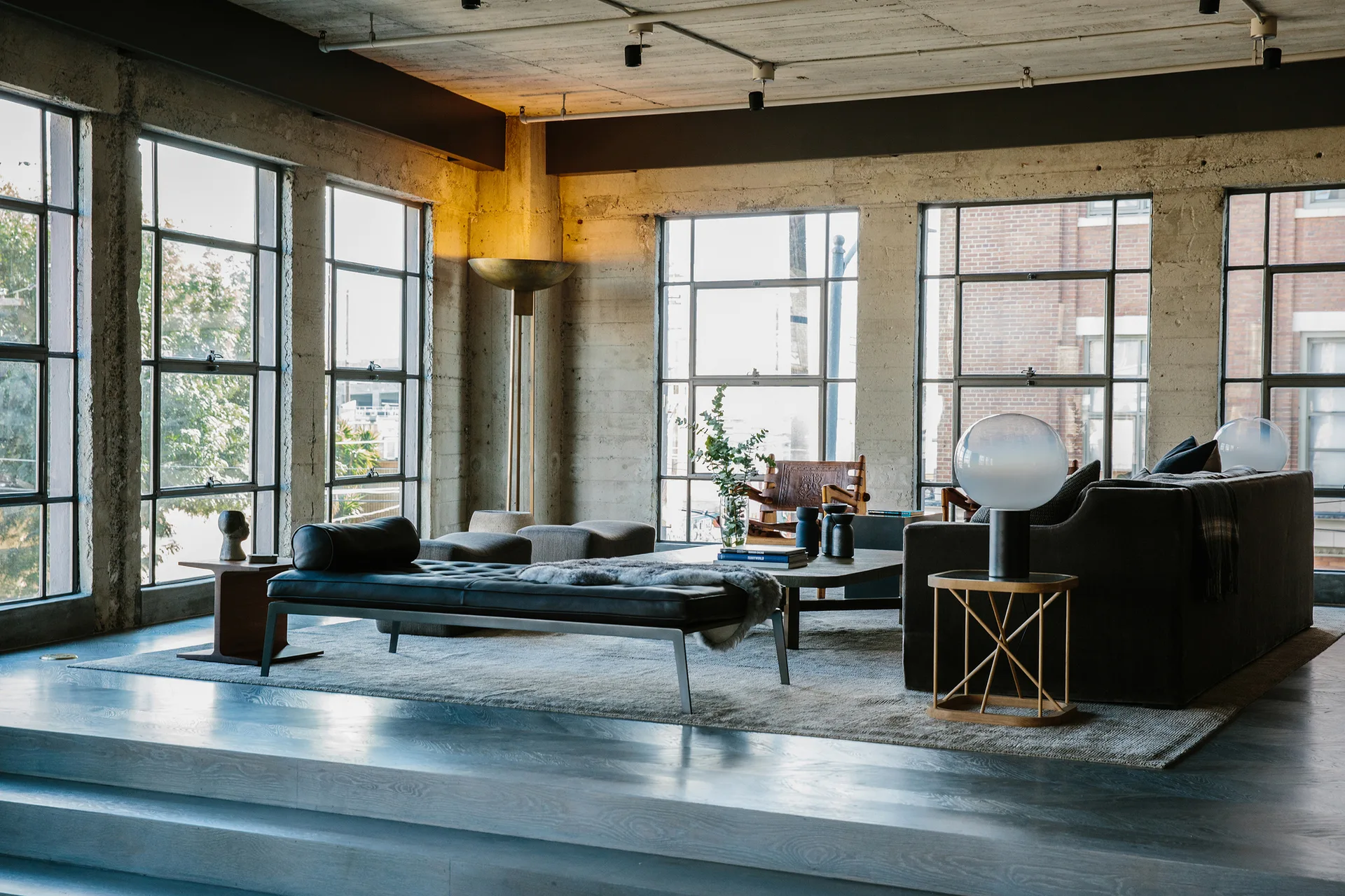 An industrial loft living room featuring a grey sofa, a black leather daybed, exposed concrete walls, and large grid-pattern windows.