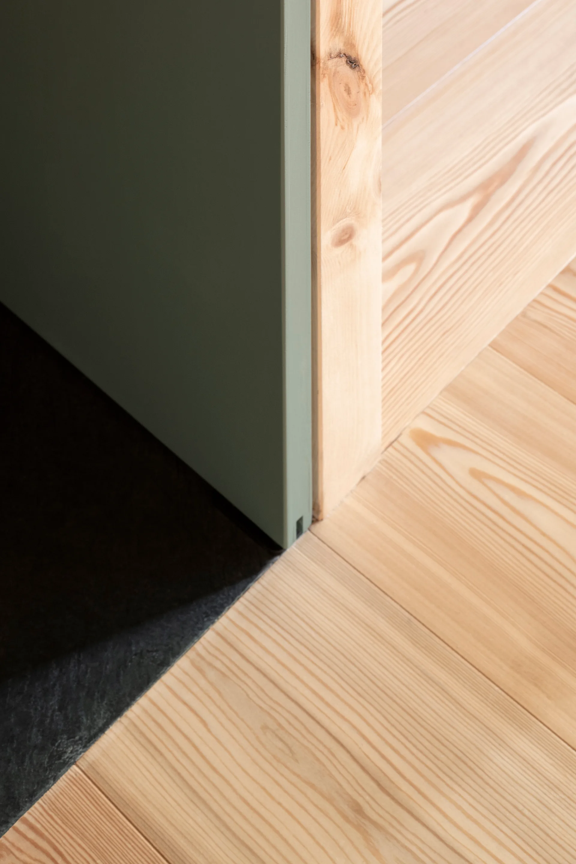 A close-up architectural detail showing a matte sage green door panel meeting light wood flooring and a vertical wood door frame.