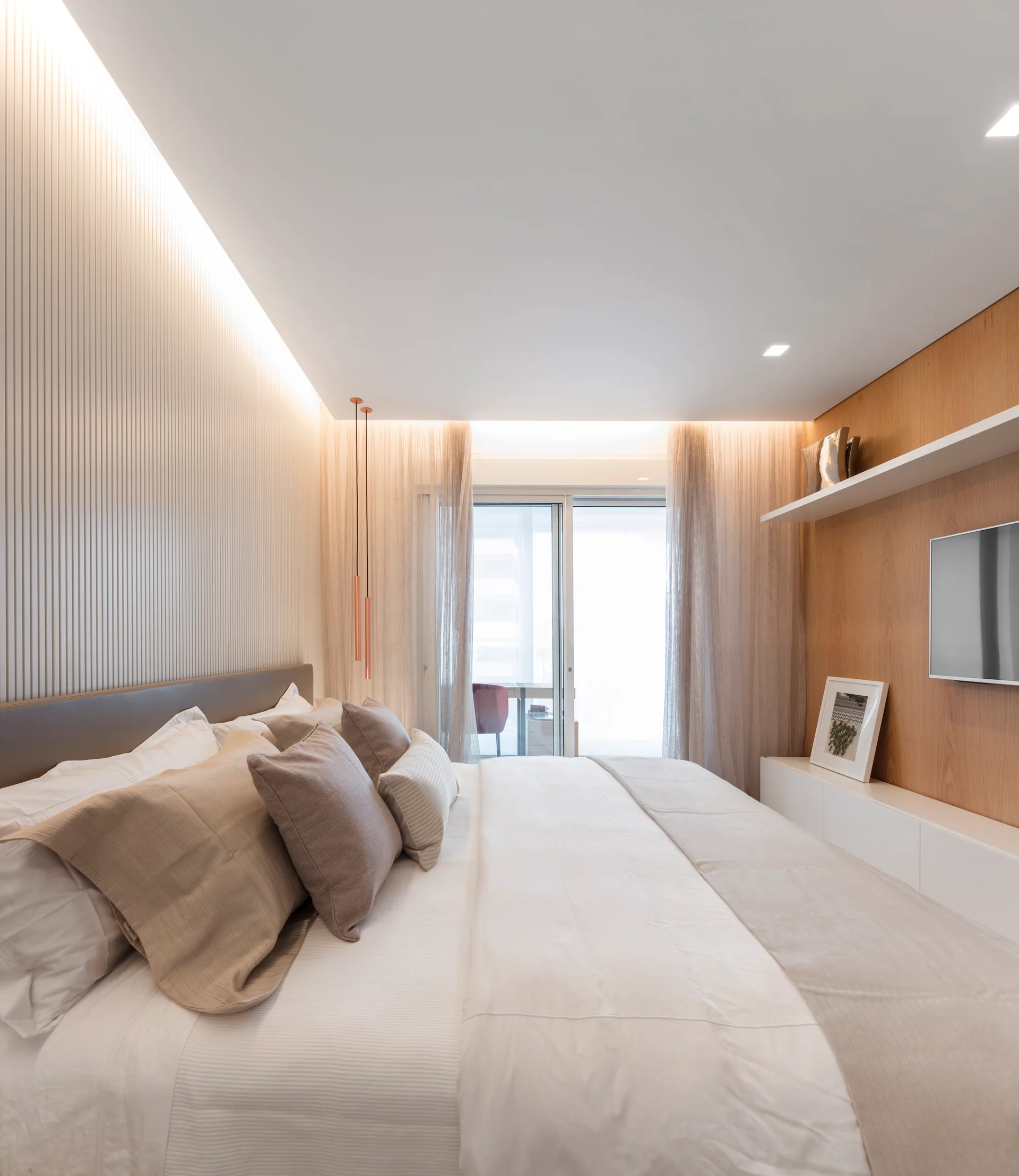 A modern bedroom featuring a bed with neutral pillows, a textured slatted accent wall, integrated ceiling cove lighting, and sheer curtains.