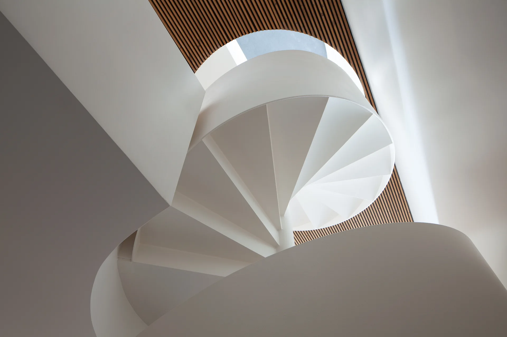 A low-angle view of a sculptural white spiral staircase rising toward a circular skylight and a slatted wood ceiling.