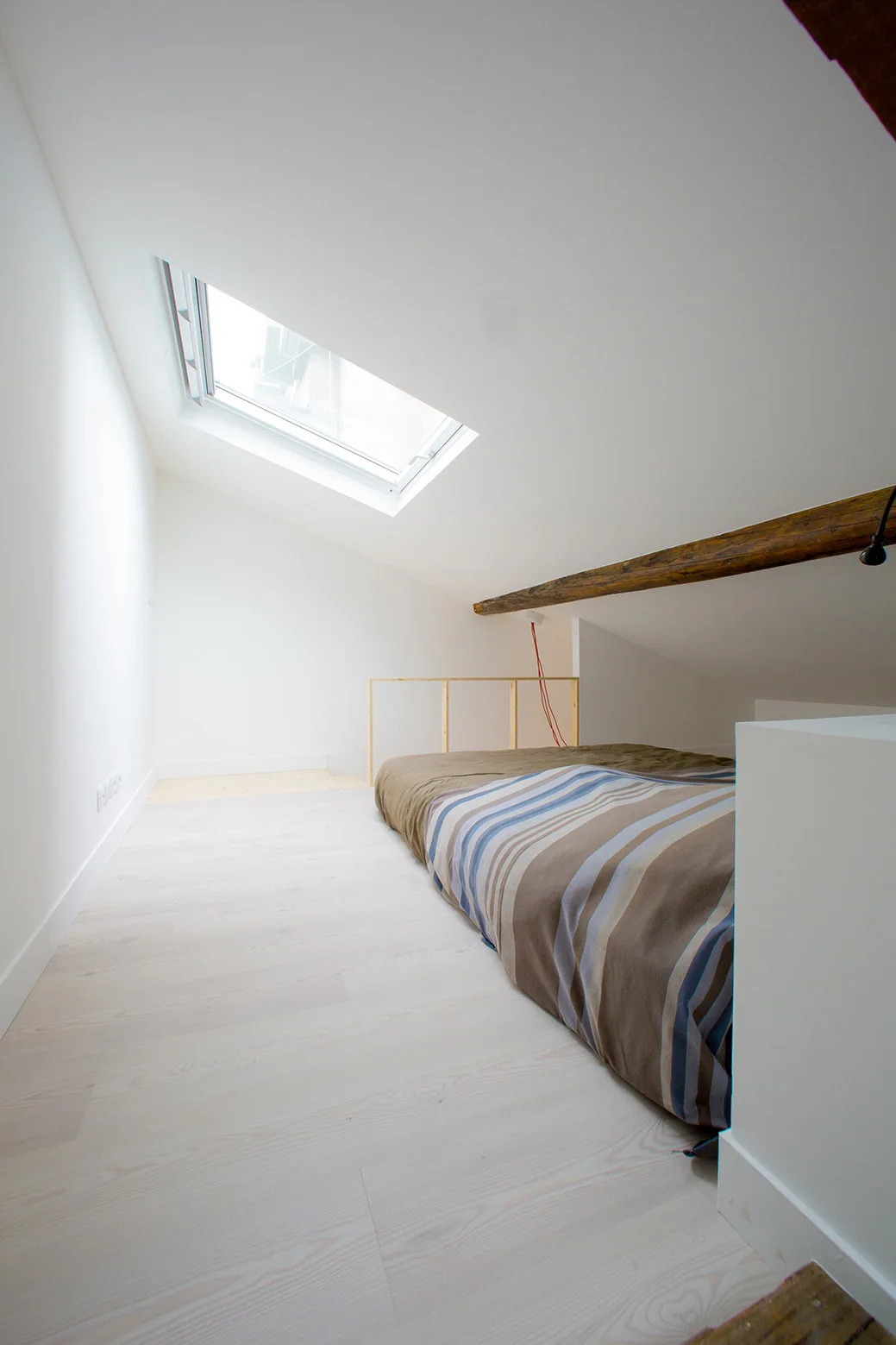 A minimalist attic loft bedroom features a low-profile bed under a sloped white ceiling with a skylight and an exposed wood beam.