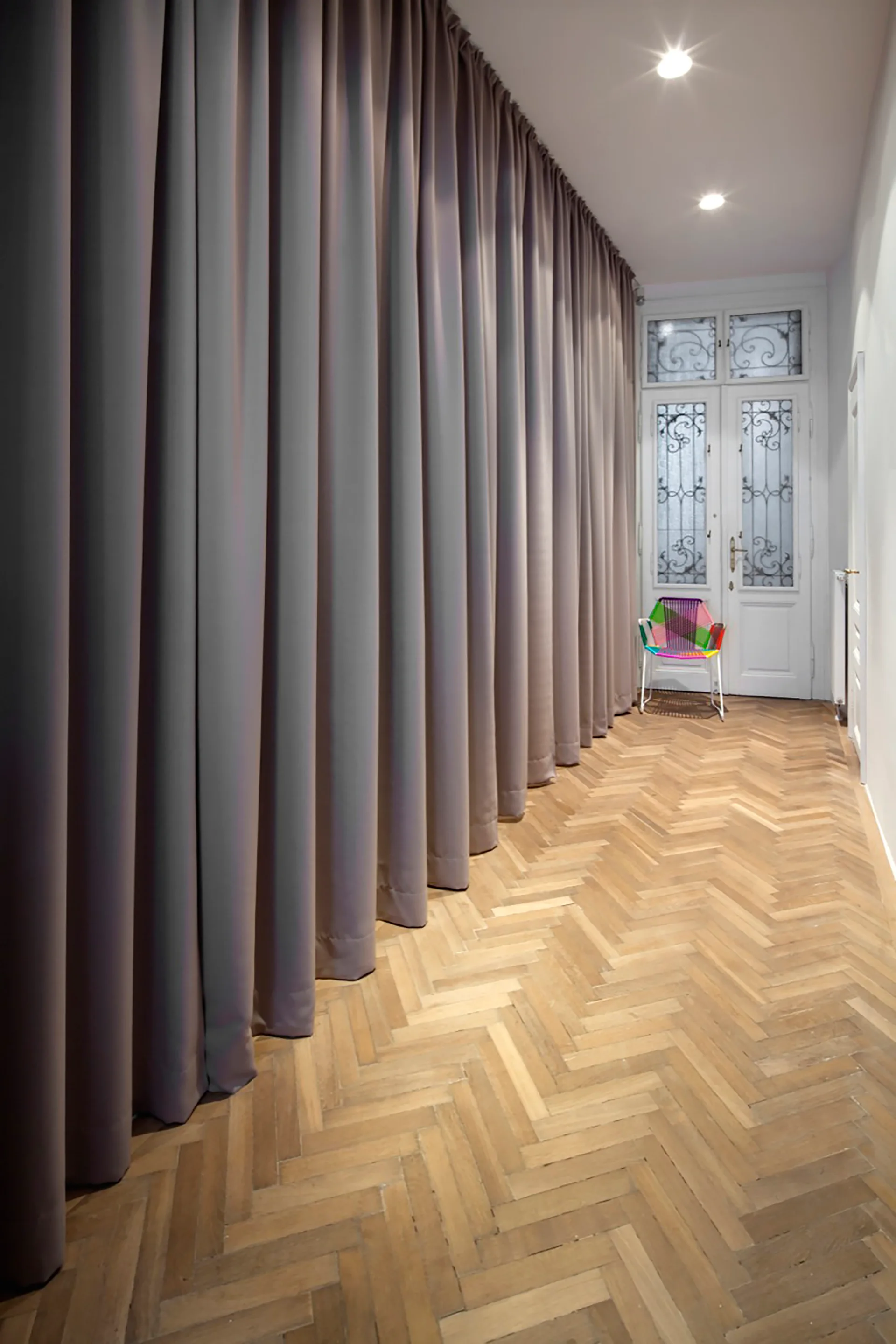 A hallway with floor-to-ceiling grey drapes on one wall, herringbone wood flooring, and a multi-colored woven chair by a decorative glass door.