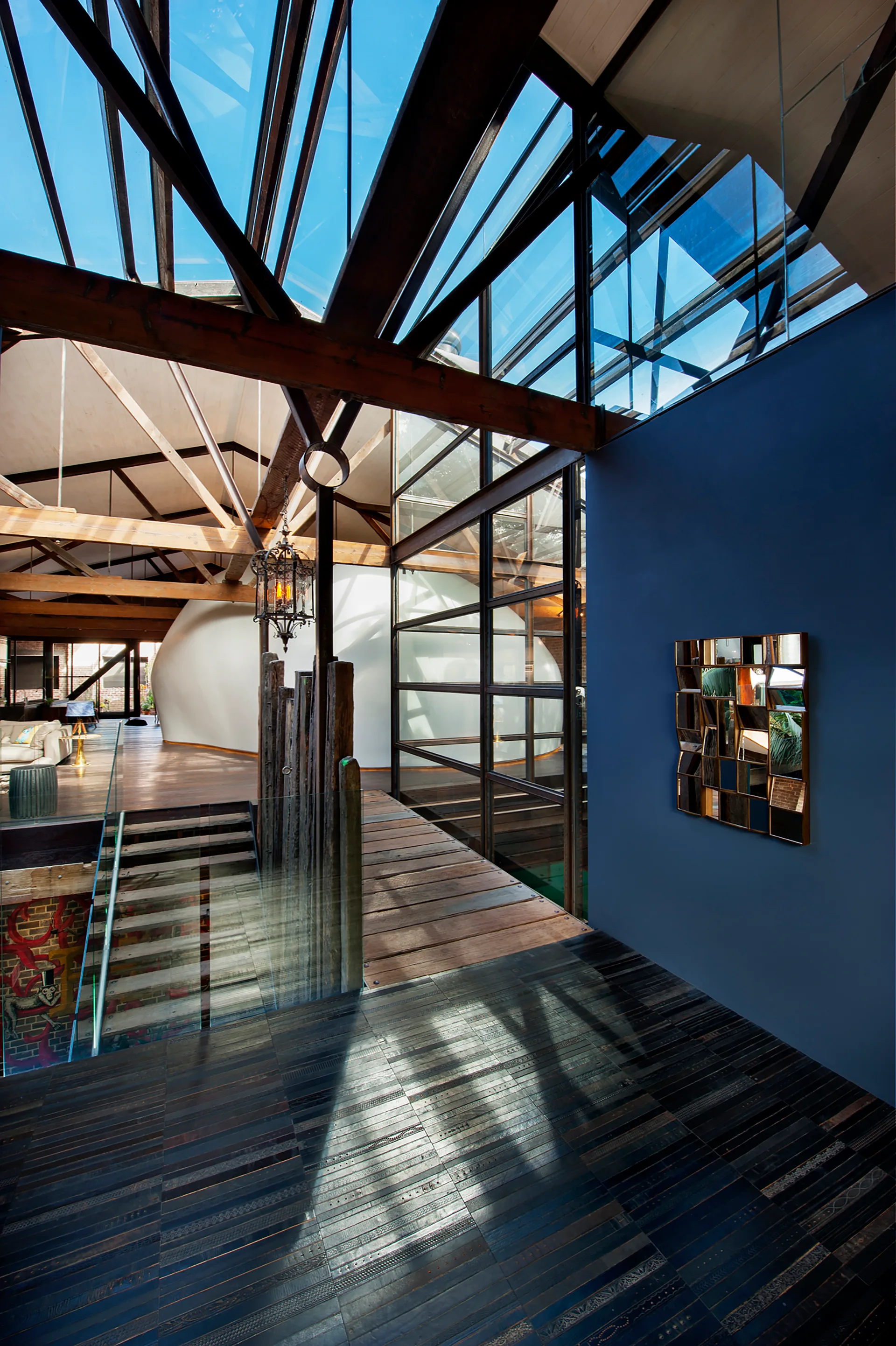 An open-concept loft hallway features a glass skylight ceiling with timber beams, a matte blue accent wall, and a geometric mirror.