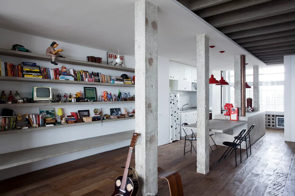 An industrial open-plan room features raw concrete structural columns, long concrete floating shelves filled with books, and a wood floor.