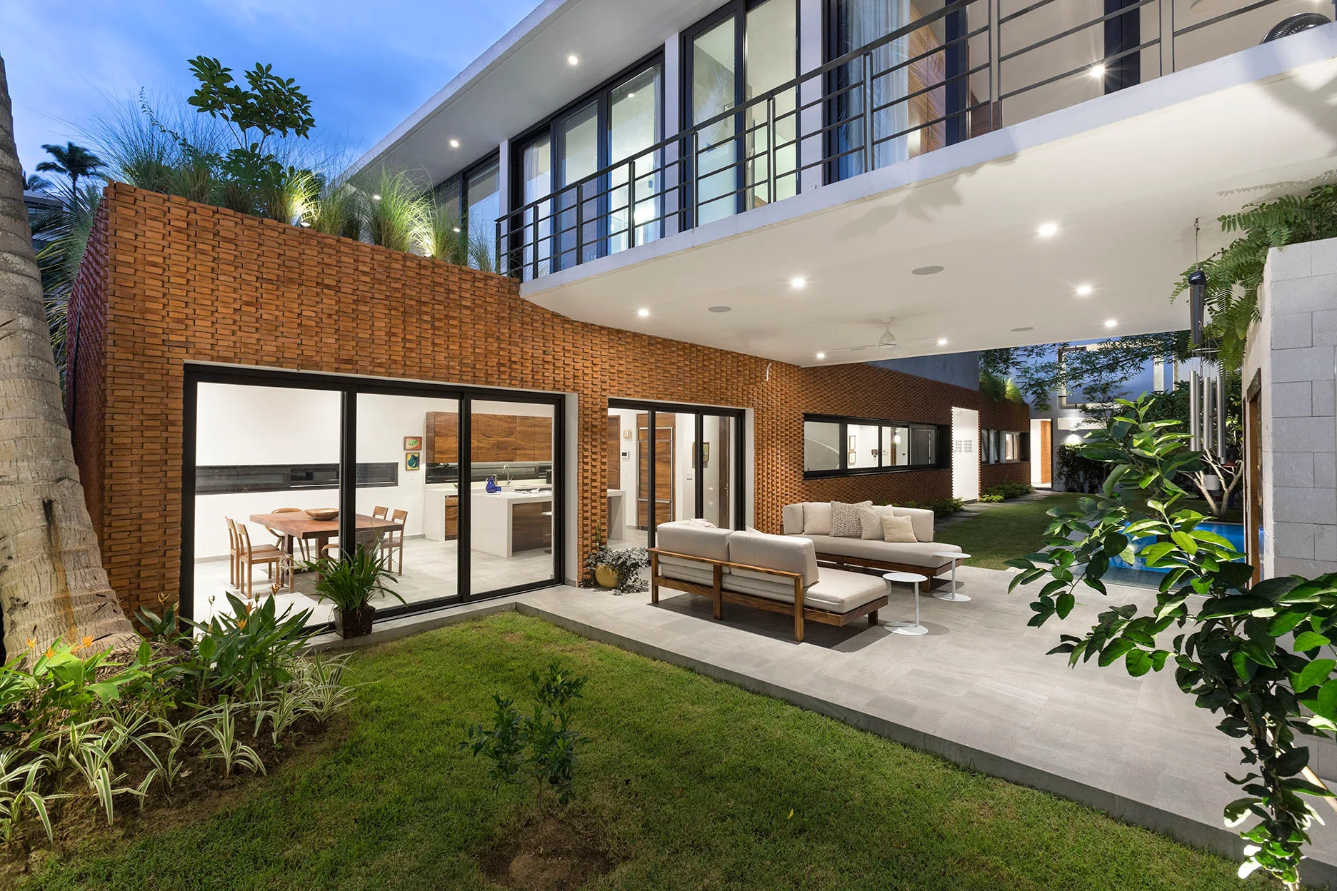A modern patio features wood-framed sofas on grey tiles, set against a terracotta brick wall with large sliding glass doors revealing the warm interior.