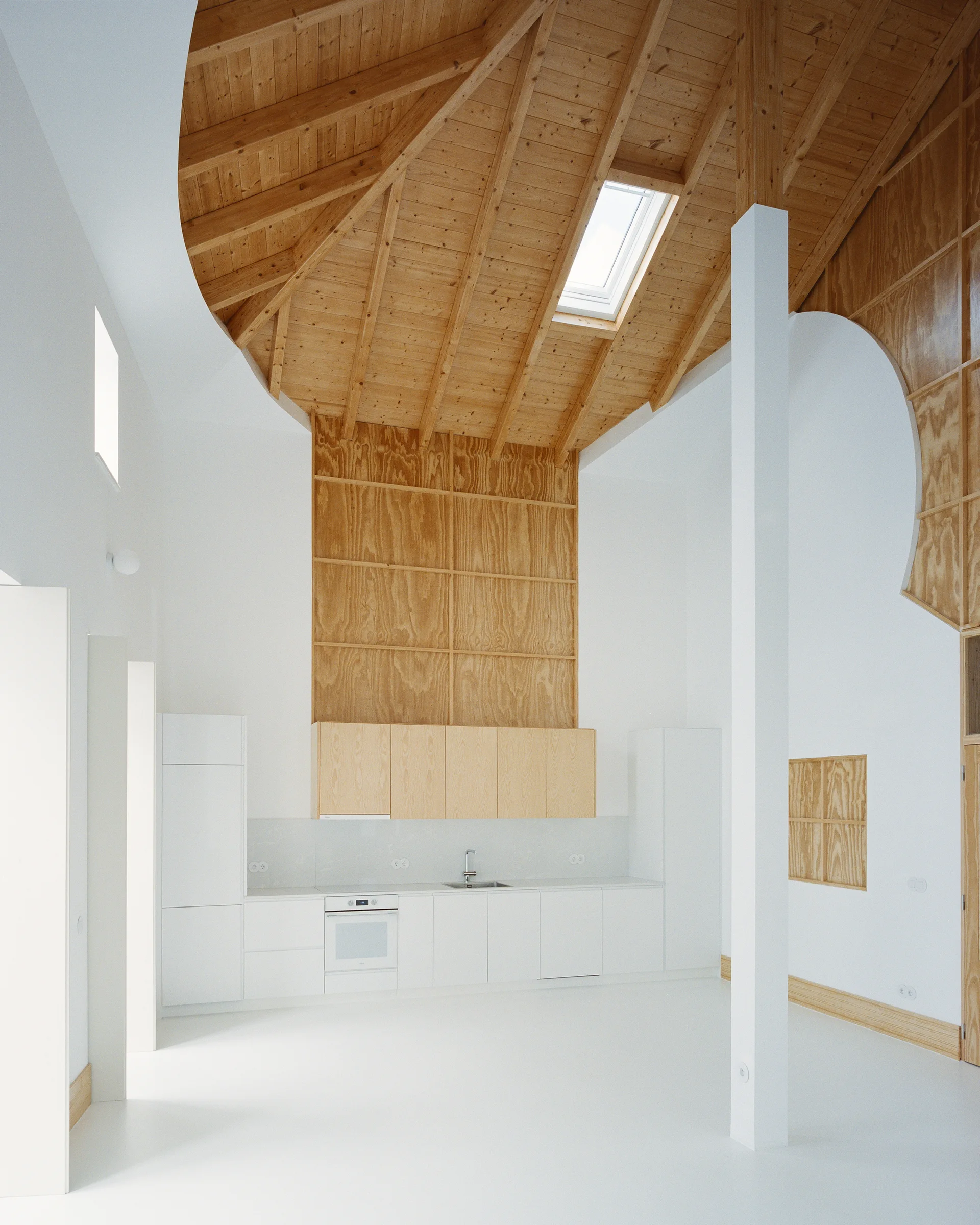 A minimalist kitchen features white base cabinets, light wood upper cabinets, a white support column, and a vaulted wood-paneled ceiling with a skylight.