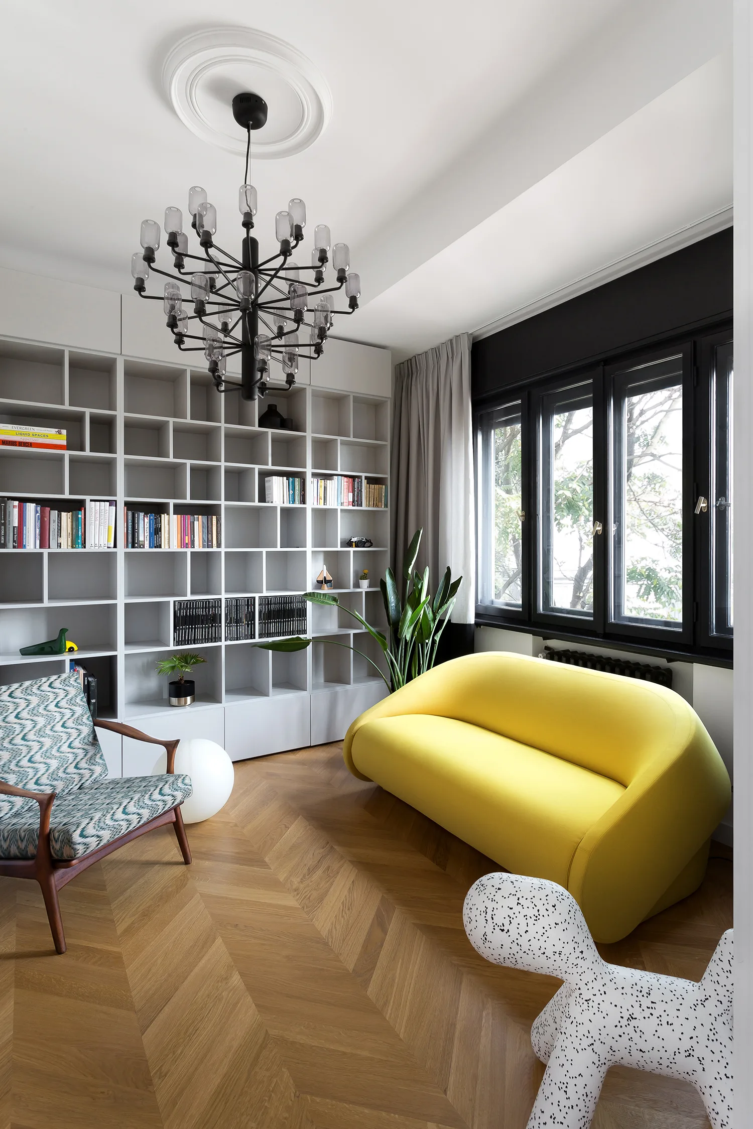 A contemporary living room featuring a bright yellow curved sofa, a large white bookshelf wall, and herringbone patterned wood floors.