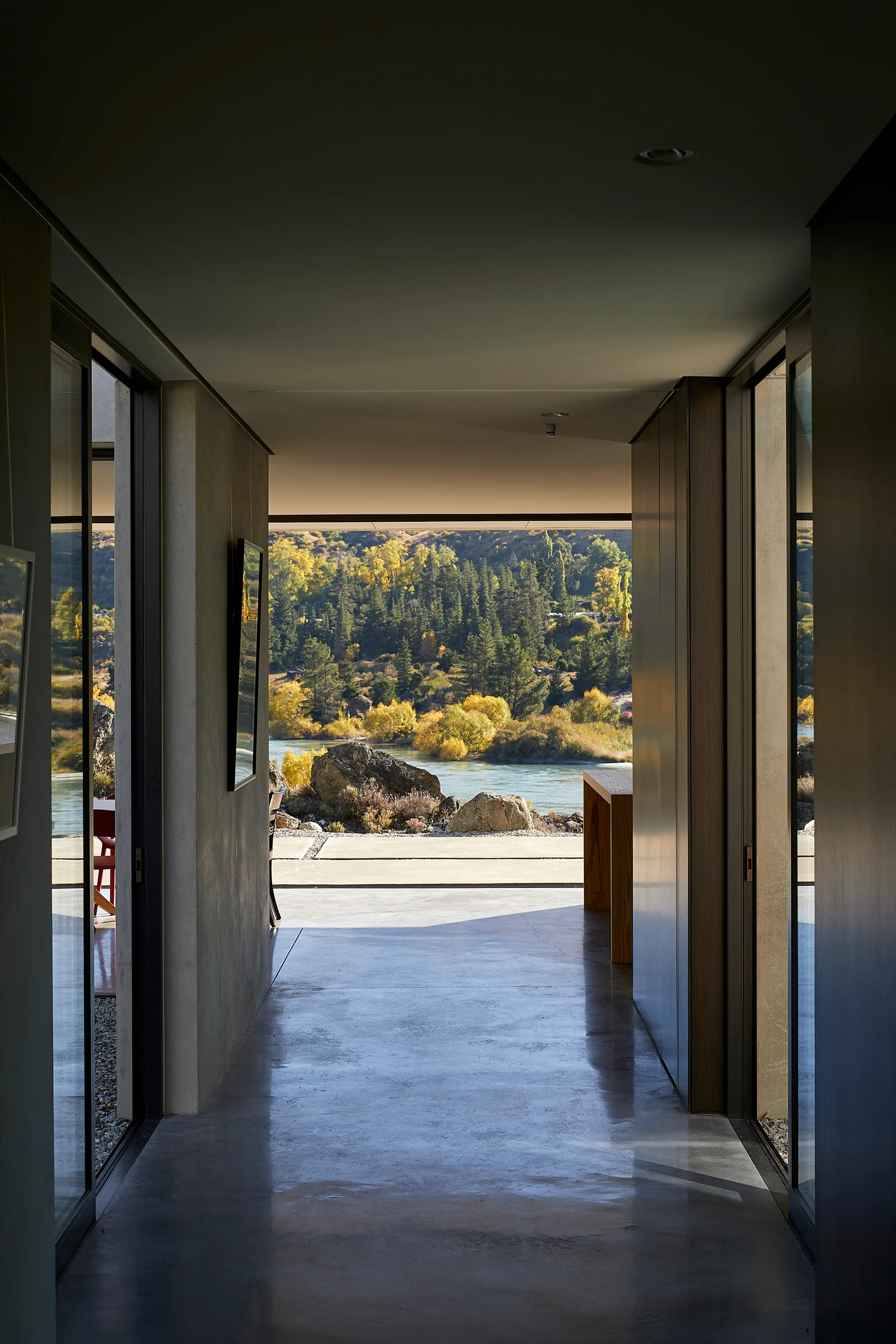 A minimalist hallway with polished concrete floors and walls looks out onto a scenic river landscape with autumn-colored trees.