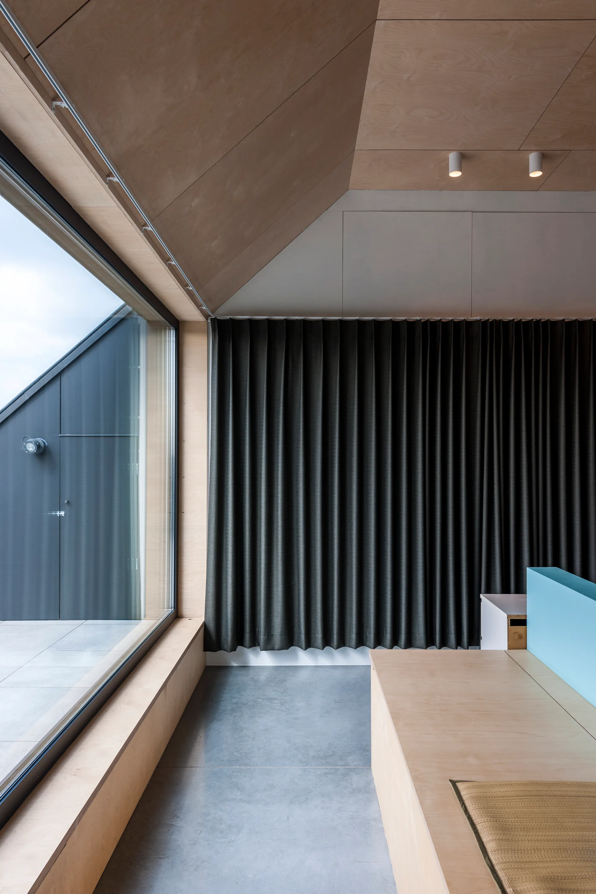 A modern minimalist room featuring an angled plywood ceiling, concrete floor, a low wooden platform bench, and dark grey pleated curtains.