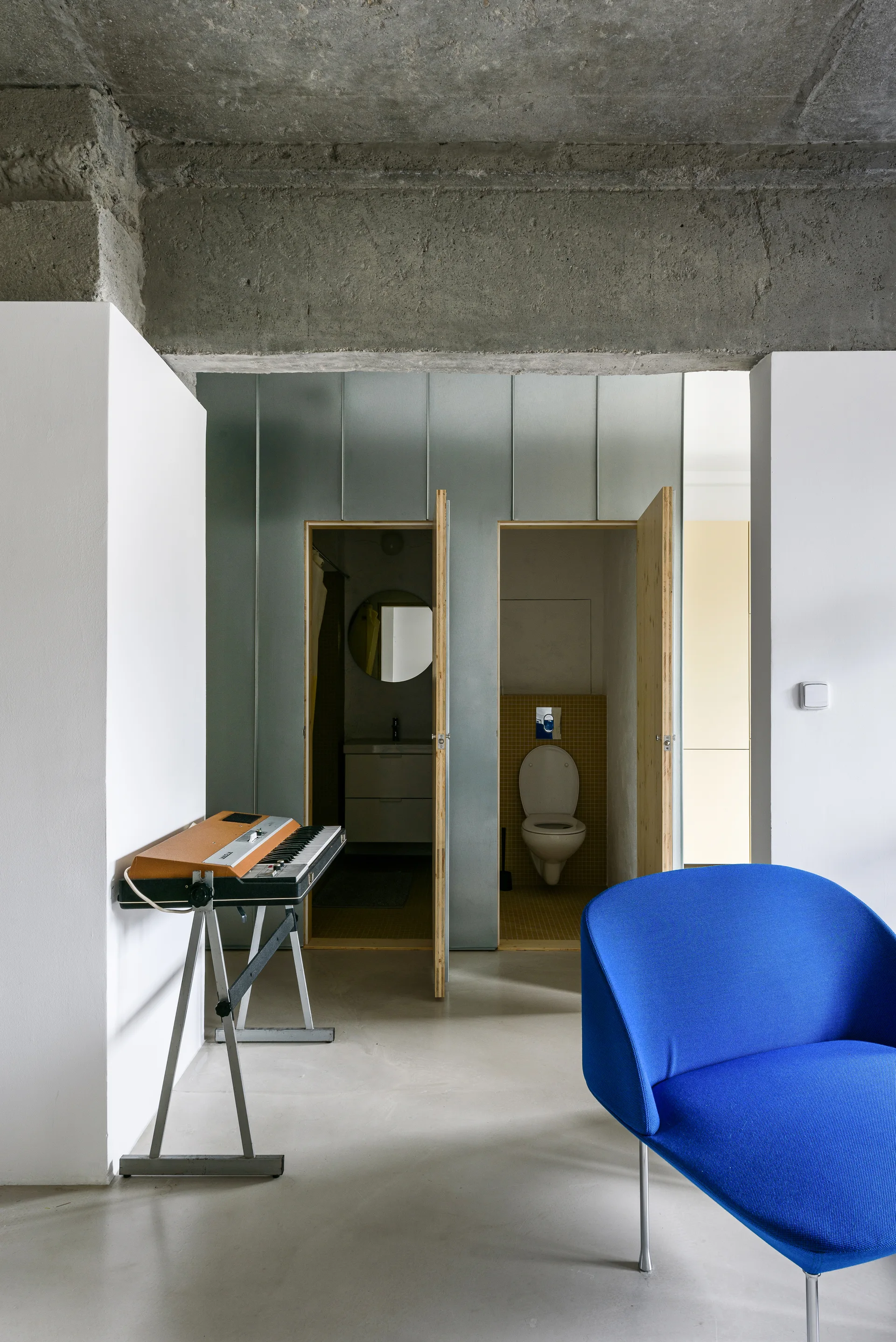 An industrial living space features an exposed concrete ceiling, a galvanized metal wall partition with open plywood doors, and a bright blue armchair.