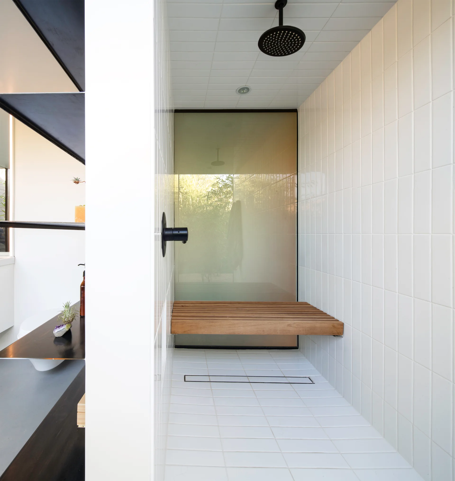 A modern walk-in shower featuring white square tile walls, a wall-mounted wooden slatted bench, black hardware, and a frosted glass backdrop.