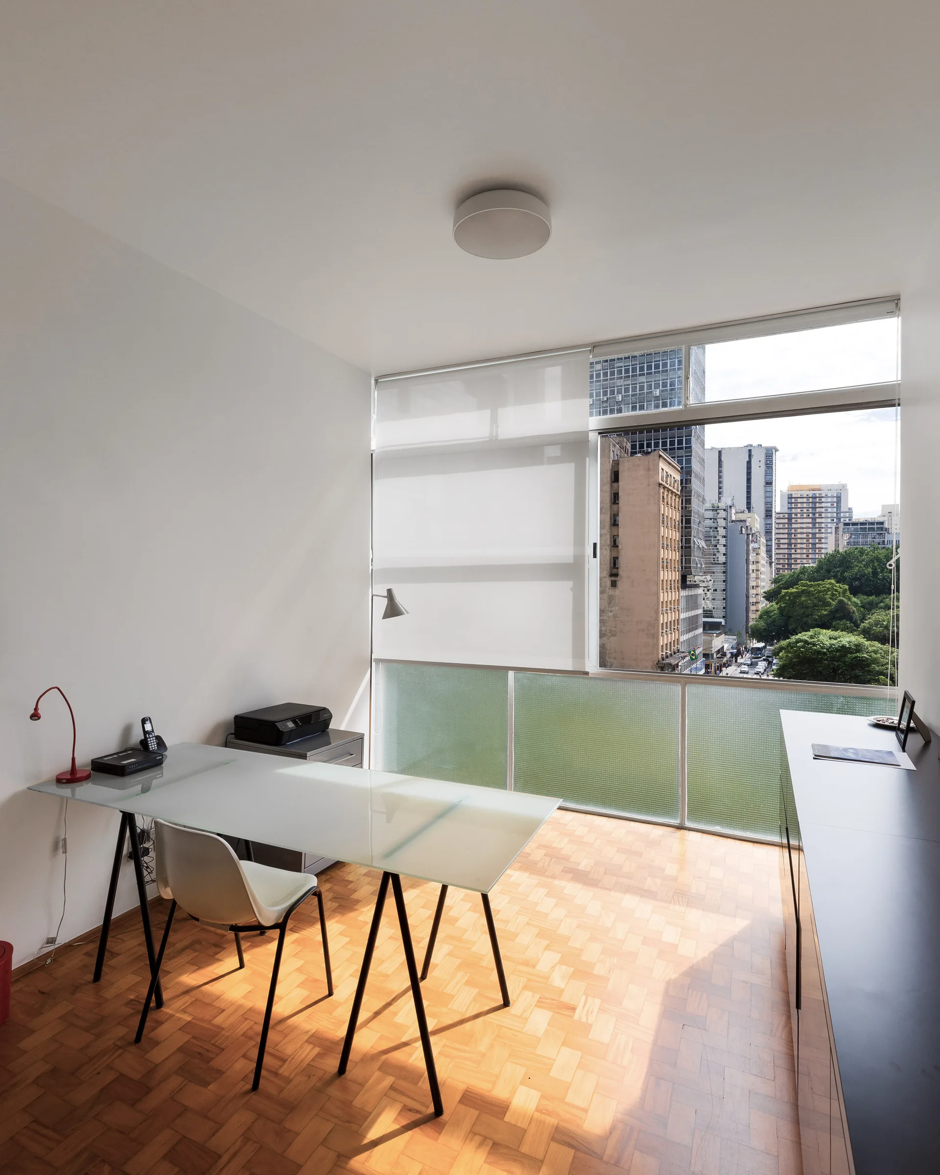 A minimalist home office features a glass-top desk, a white shell chair, warm parquet wood flooring, and a large window overlooking a cityscape.