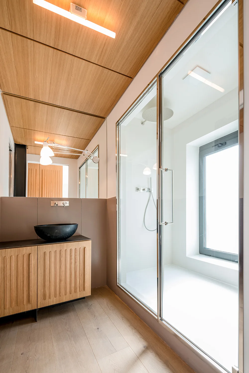 A modern bathroom showing a fluted wood vanity with a black vessel sink and a glass-enclosed shower next to it under a wood plank ceiling.