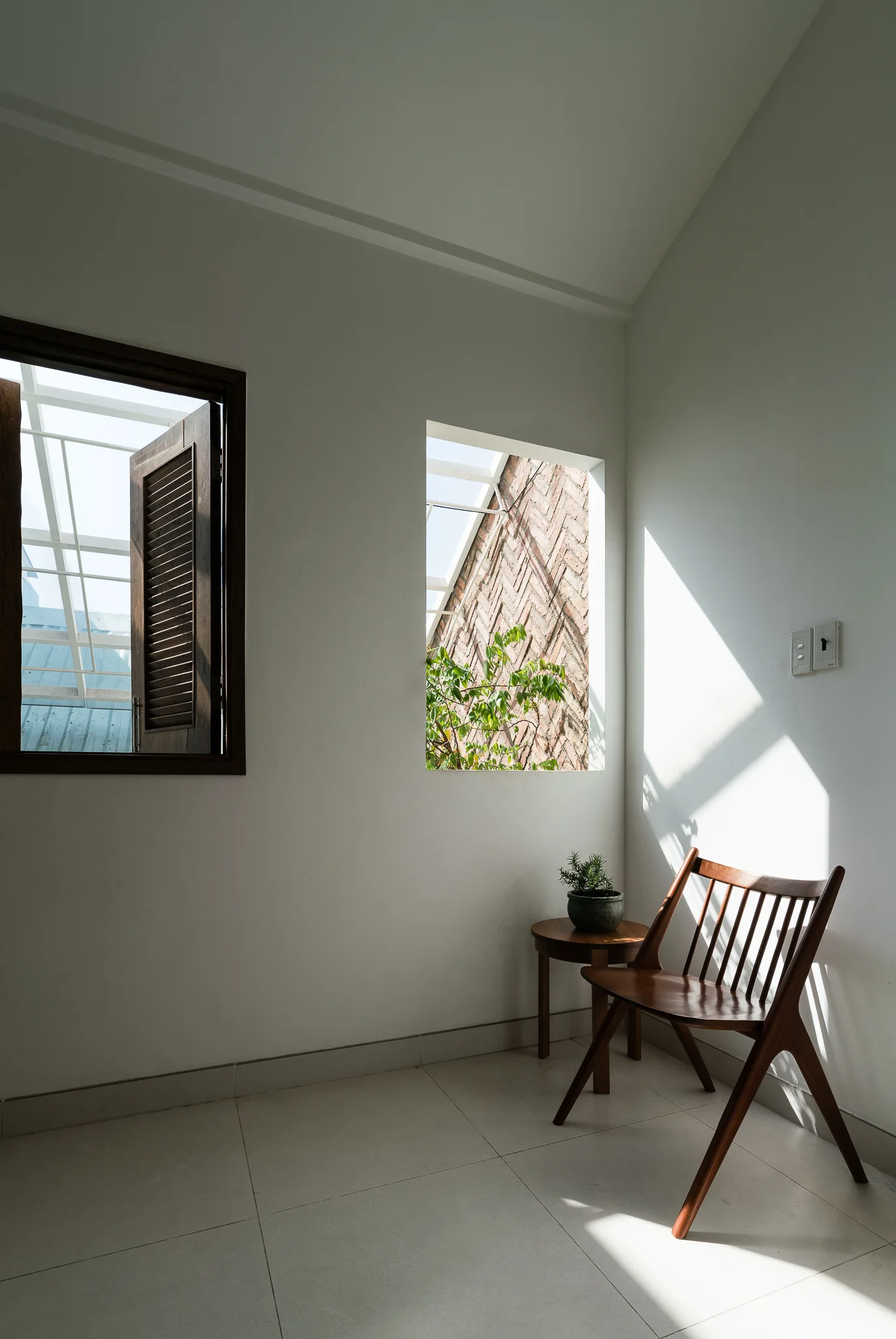 A minimalist white room corner features a wooden spindle chair next to a small side table and a window framing a view of a brick wall.