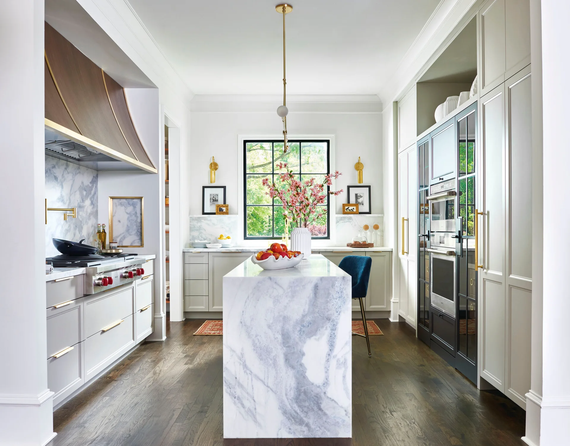 A bright kitchen features a white marble waterfall island, custom white cabinets with brass hardware, a dark wood floor, and a black-framed window.