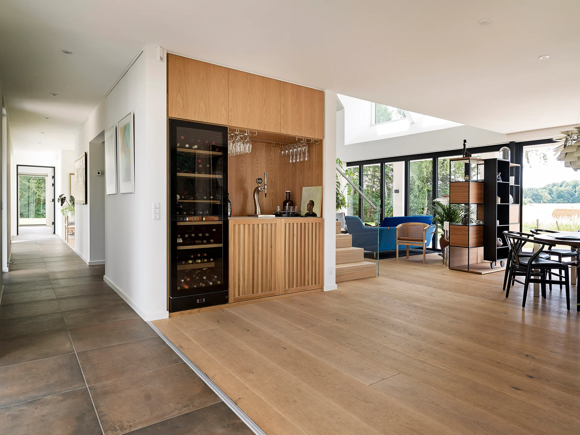 A modern open-concept living space featuring a custom wooden built-in bar with a wine cooler, light wood floors, and a blue sofa in the background.