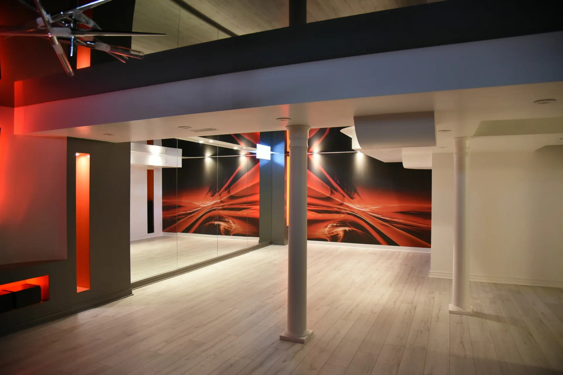 A modern home studio space features light wood floors, white structural pillars, a large mirrored wall, and an abstract red and orange graphic mural.