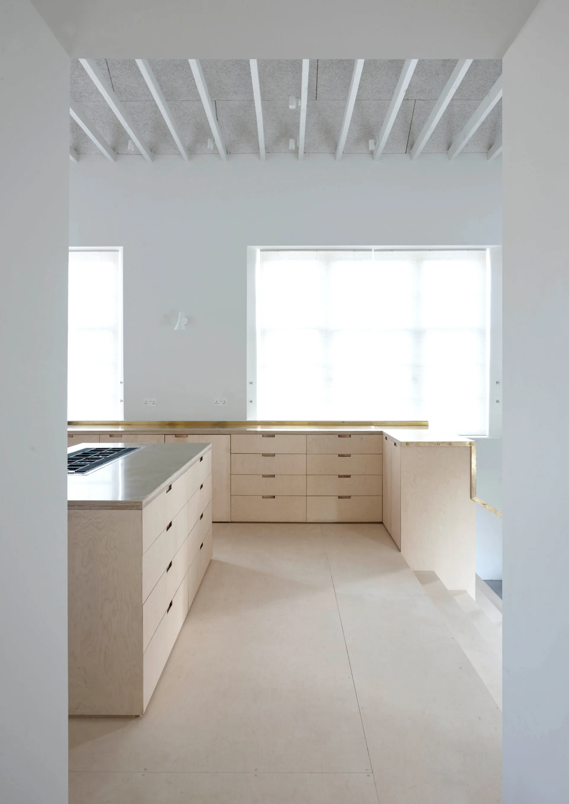 A minimalist kitchen features custom light plywood cabinetry, a matching island with an integrated stove, and white exposed ceiling beams.
