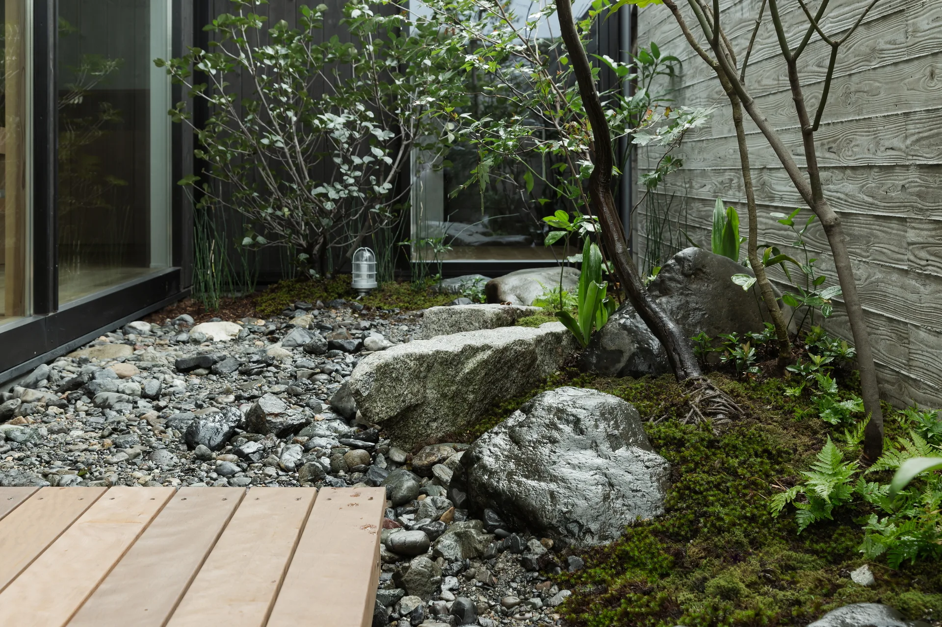 A minimalist courtyard garden features a mossy stone arrangement, river pebbles, a textured board-formed concrete wall, and a wooden deck.