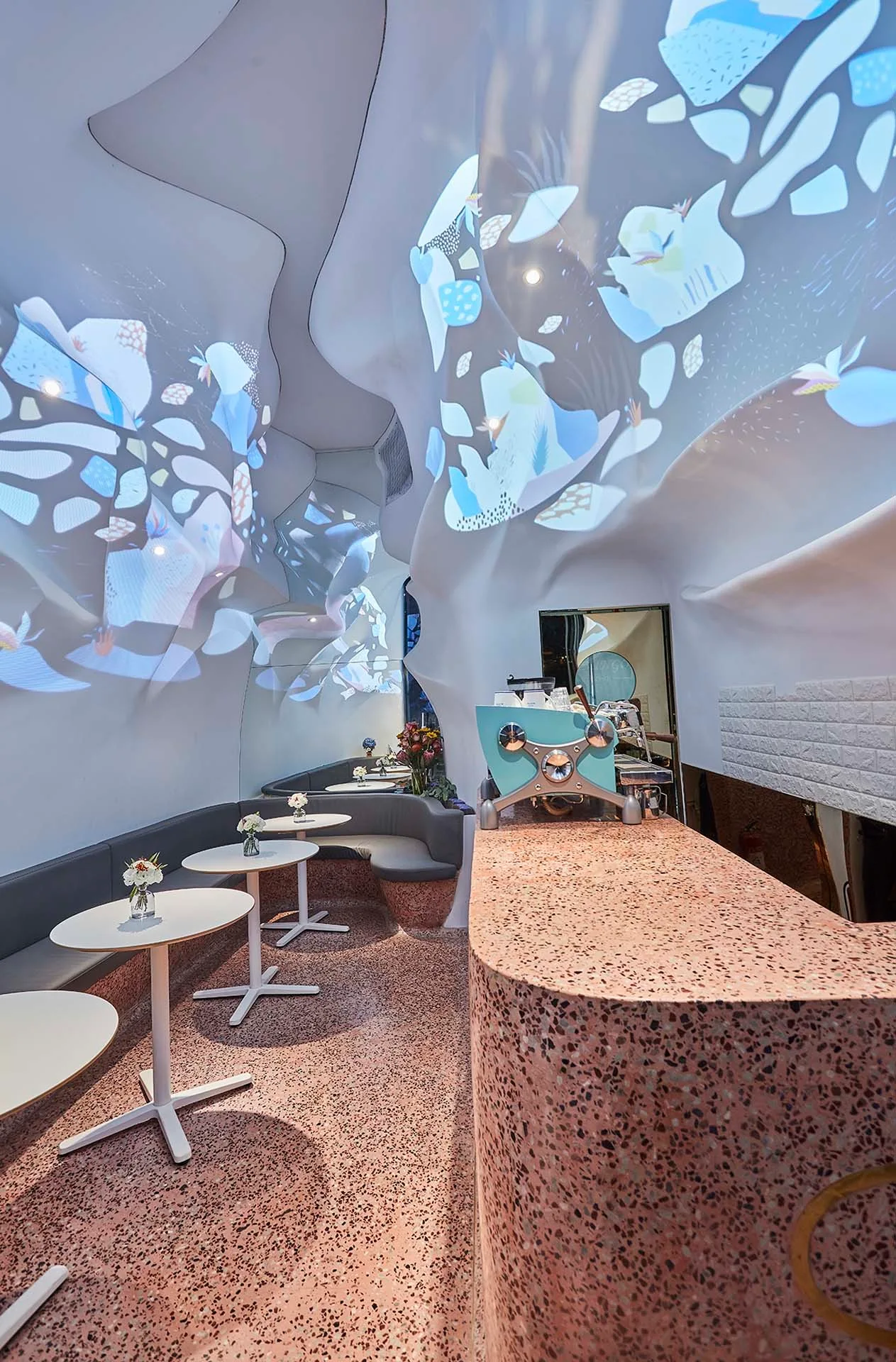 A modern cafe interior featuring curved sculptural white walls with projected light patterns, small bistro tables, and a pink terrazzo counter.