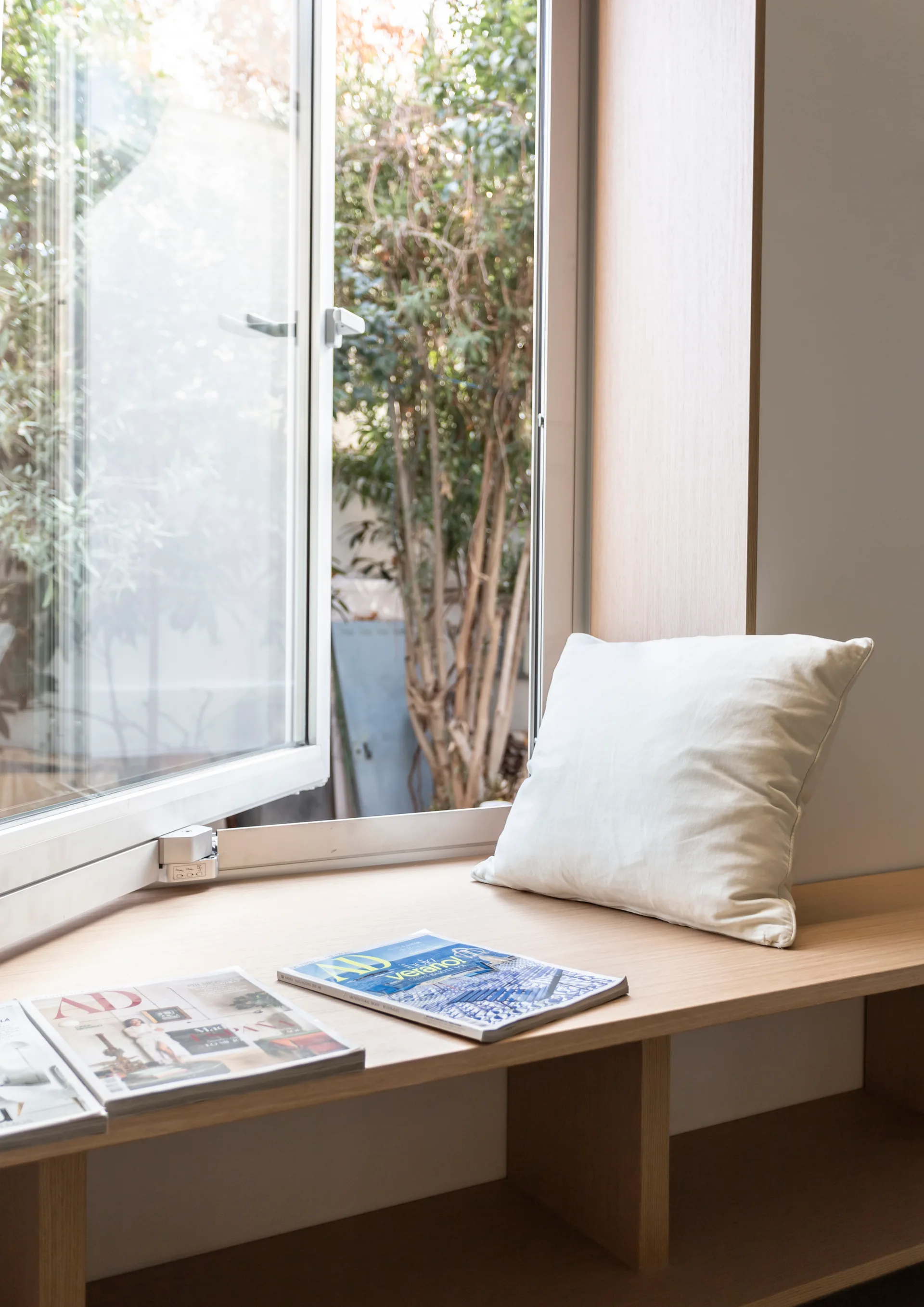 A light wood built-in window bench holds a white square cushion and magazines next to an open glass casement window.