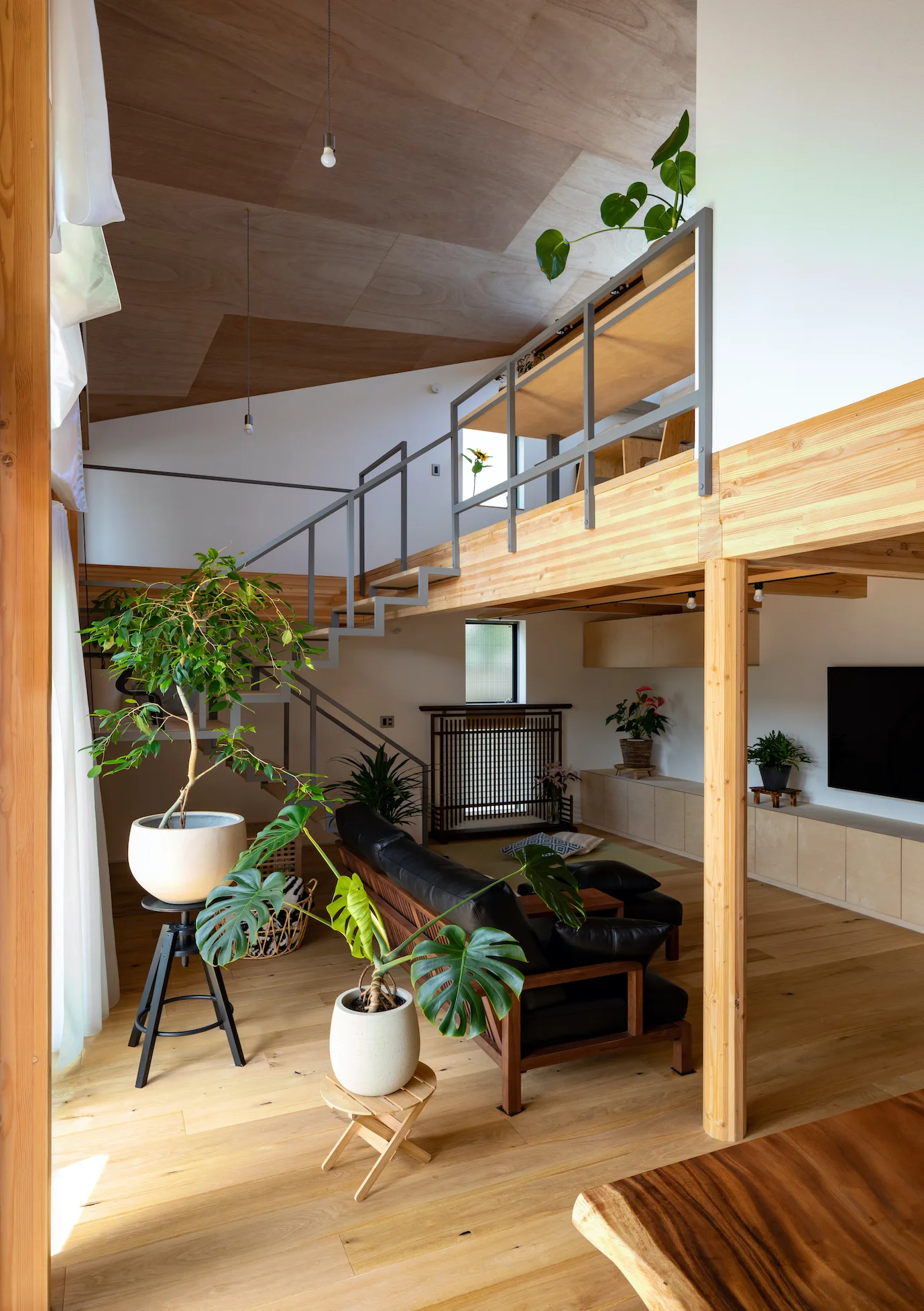 A split-level living room features a black leather sofa, light wood floors, potted plants, and an open metal staircase leading to a mezzanine.