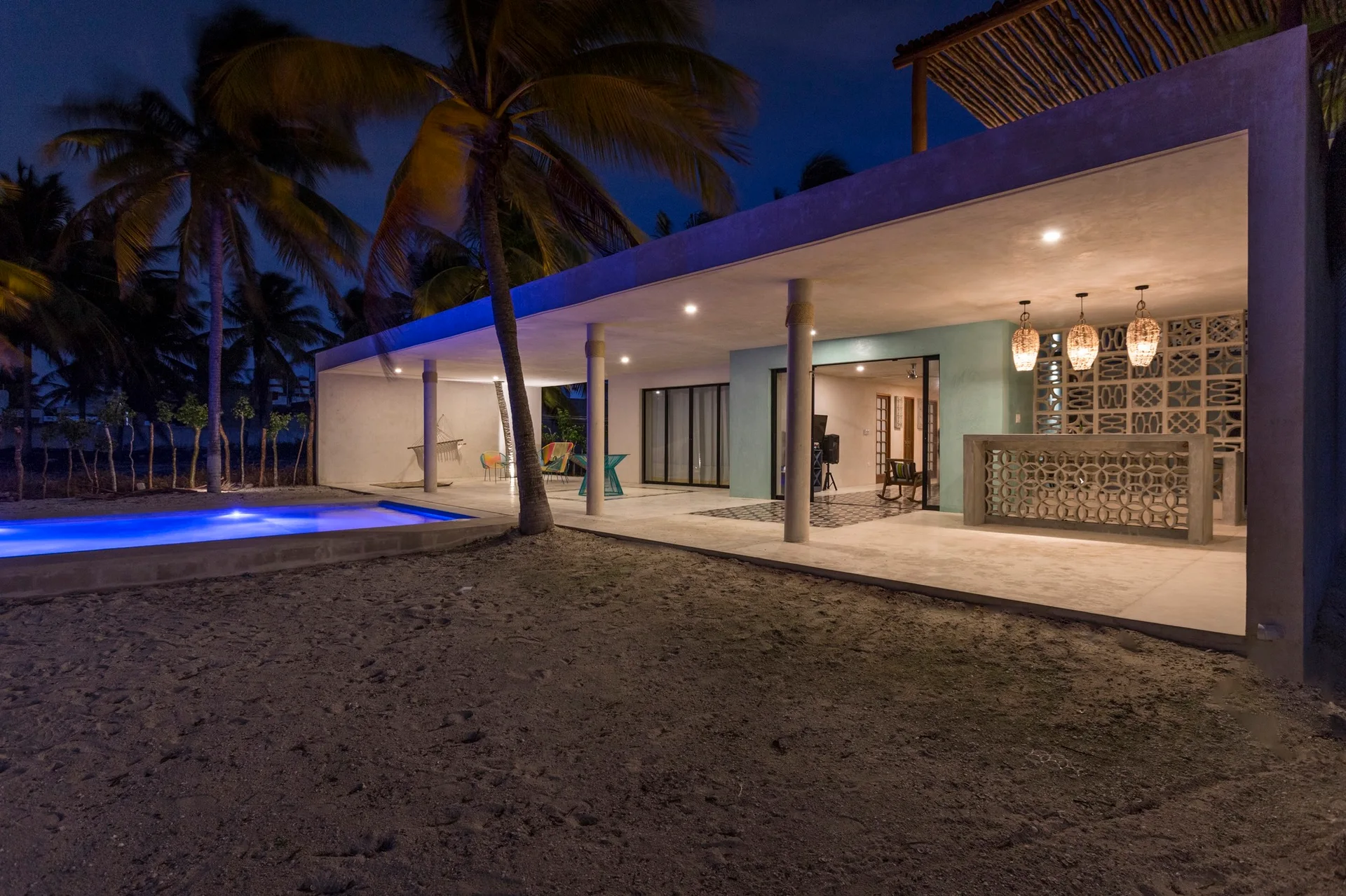 An illuminated open-air concrete patio at night featuring a swimming pool, turquoise accent wall, and a bar with decorative breeze blocks.