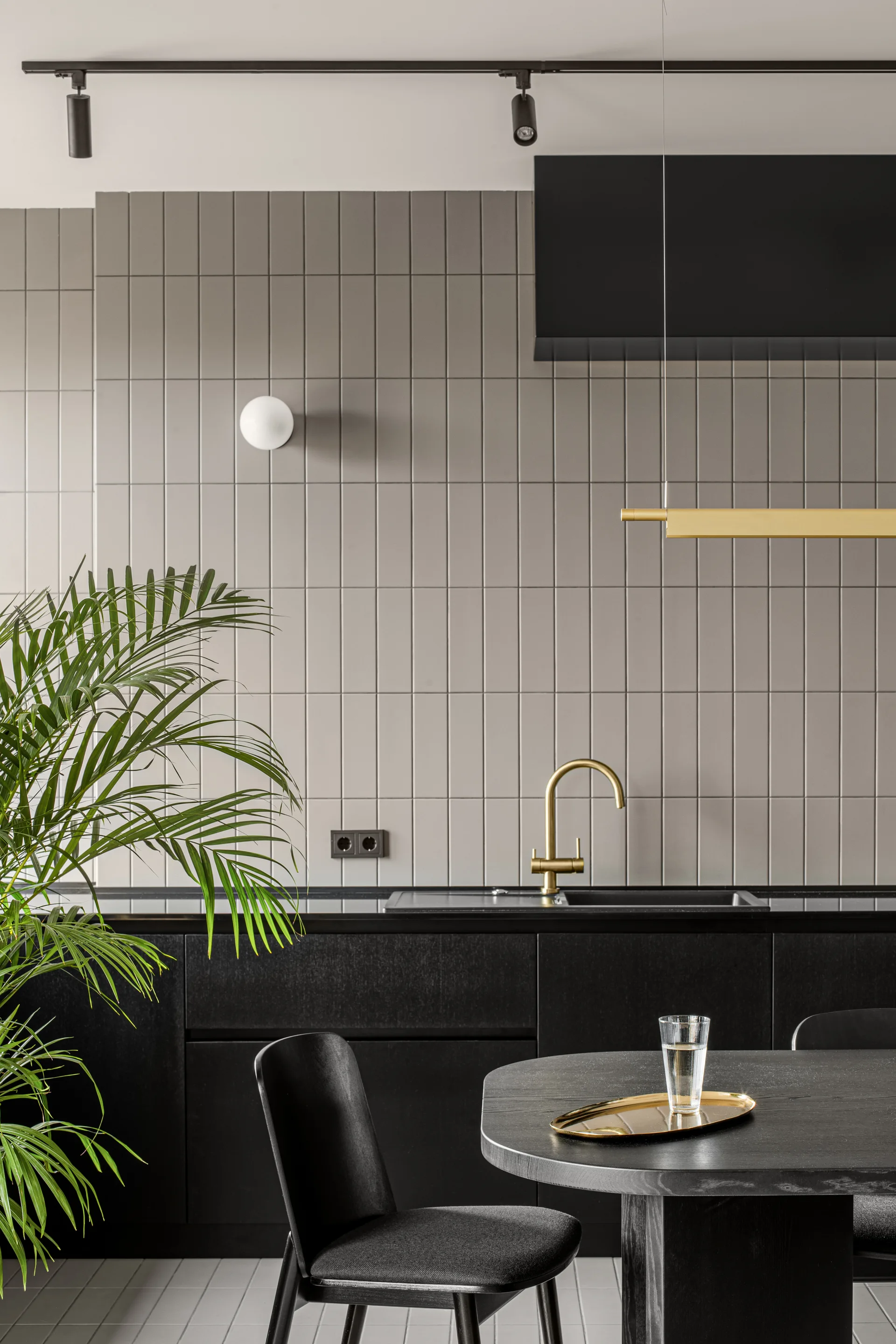 A modern kitchen featuring matte black lower cabinets, a vertical grey tile backsplash, a gold faucet, and a black dining set.