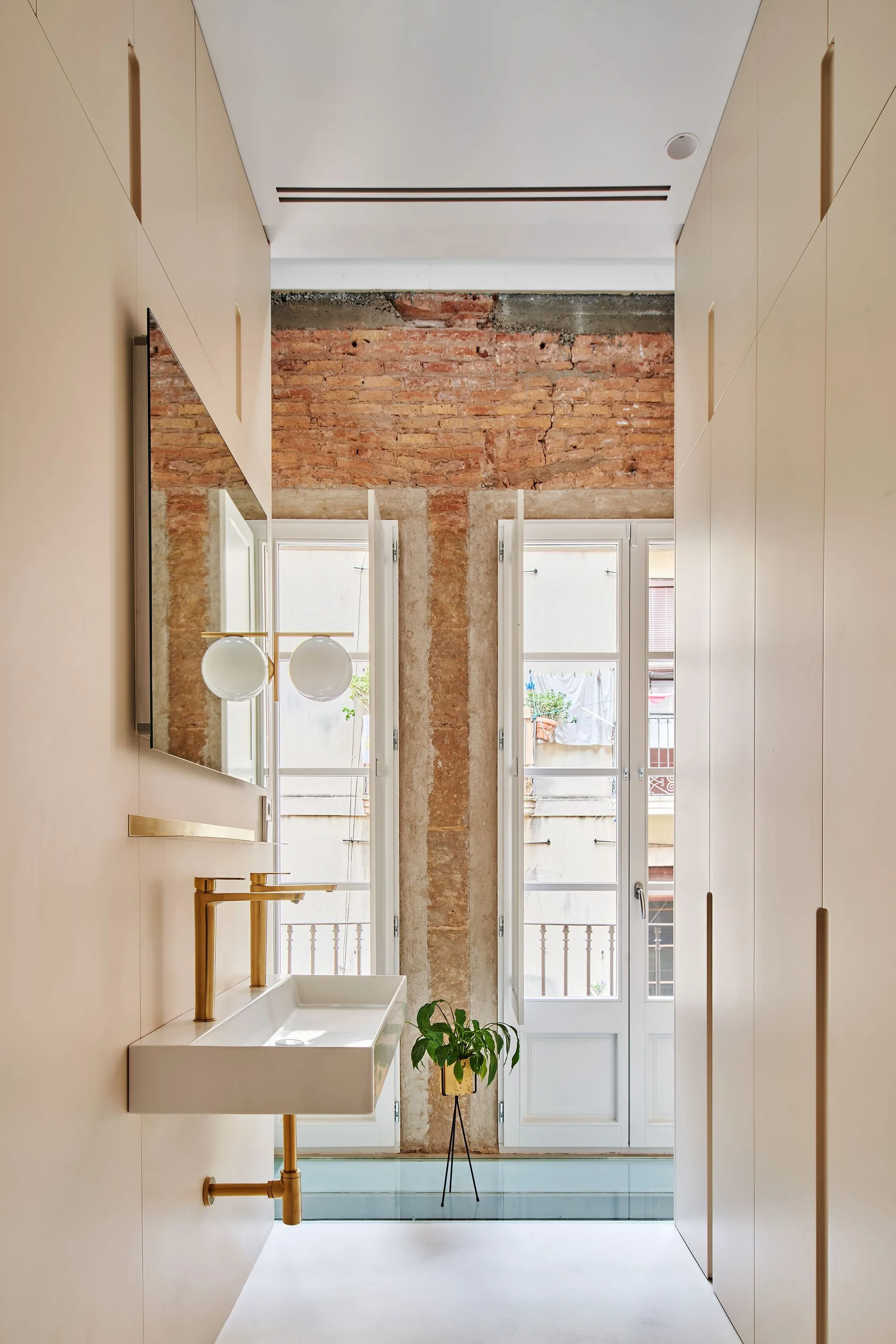 A narrow bathroom features matte beige integrated cabinets, a wall-mounted sink with gold hardware, and tall windows flanked by exposed brick.