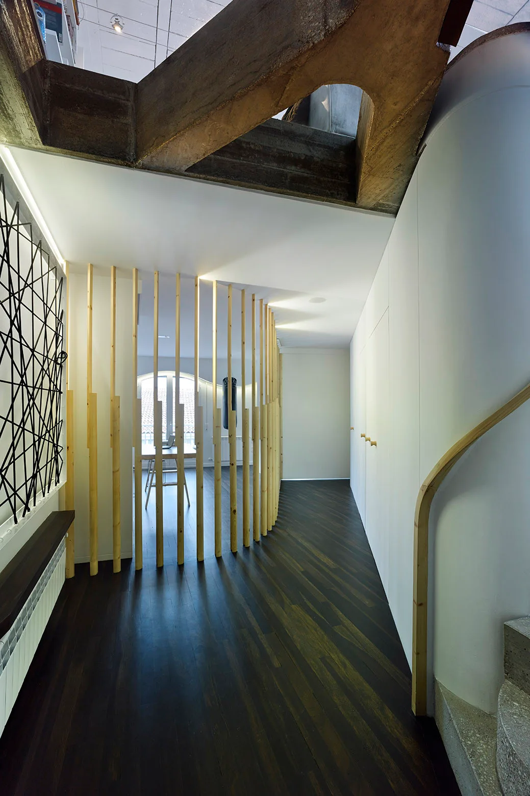 A modern hallway features a curved light wood slat screen separating a room, dark wood floors, and a seamless white storage wall.