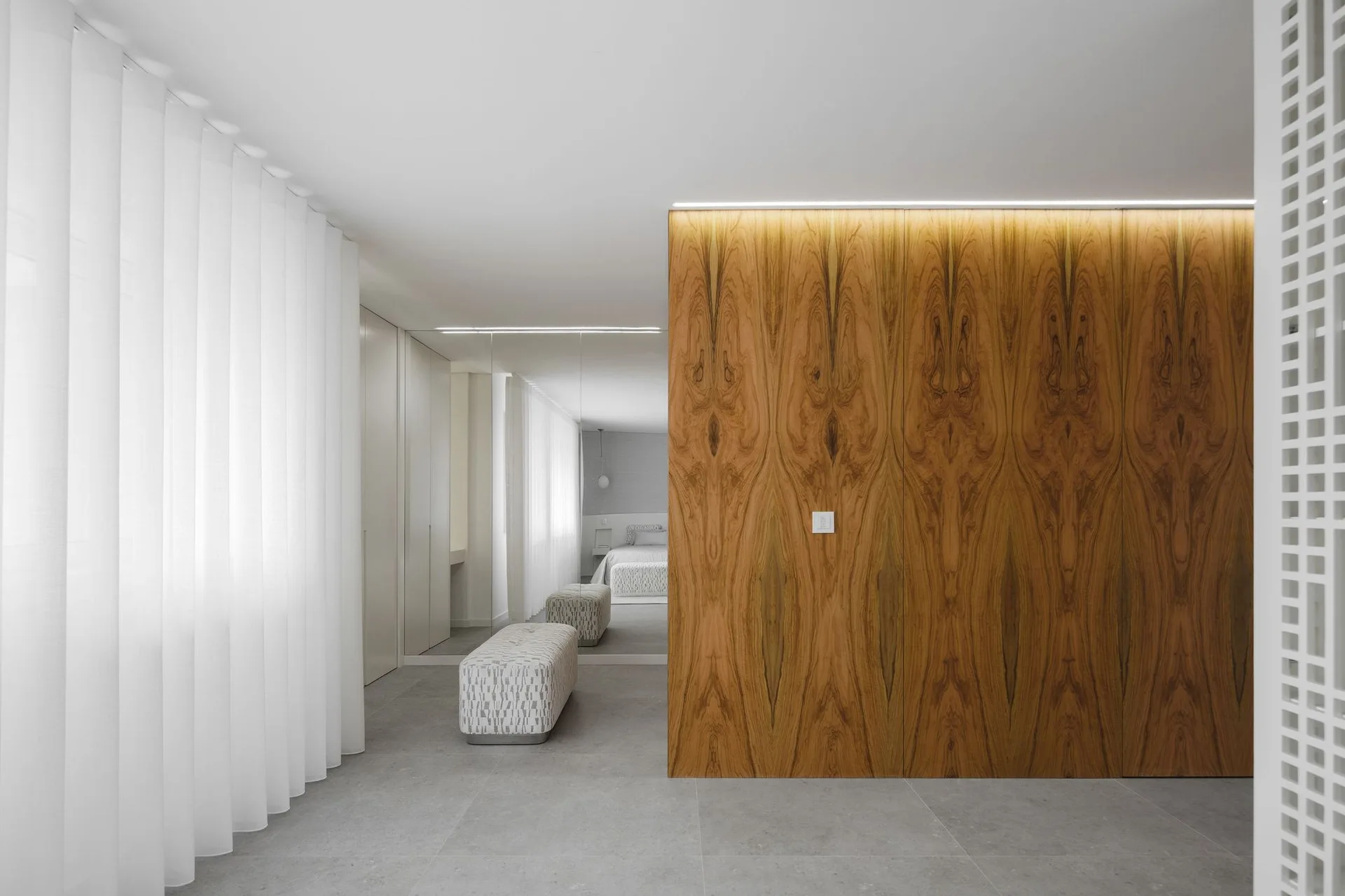 A minimalist dressing area features a bookmatched wood accent wall, a patterned upholstered bench, and a large mirror reflecting the bed.