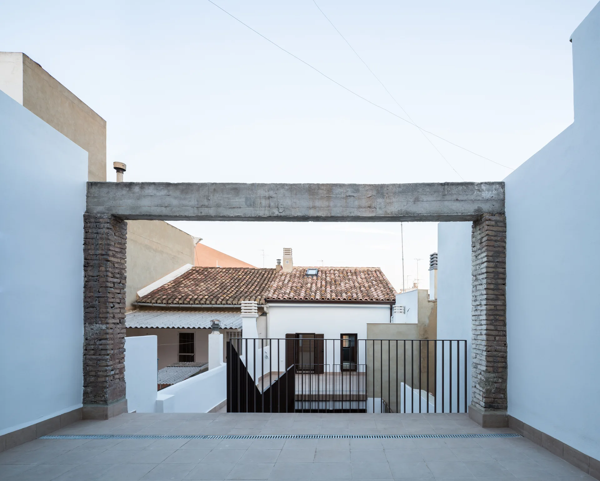 A paved outdoor terrace framed by two white walls, brick pillars, and a raw concrete overhead beam overlooking residential rooftops.