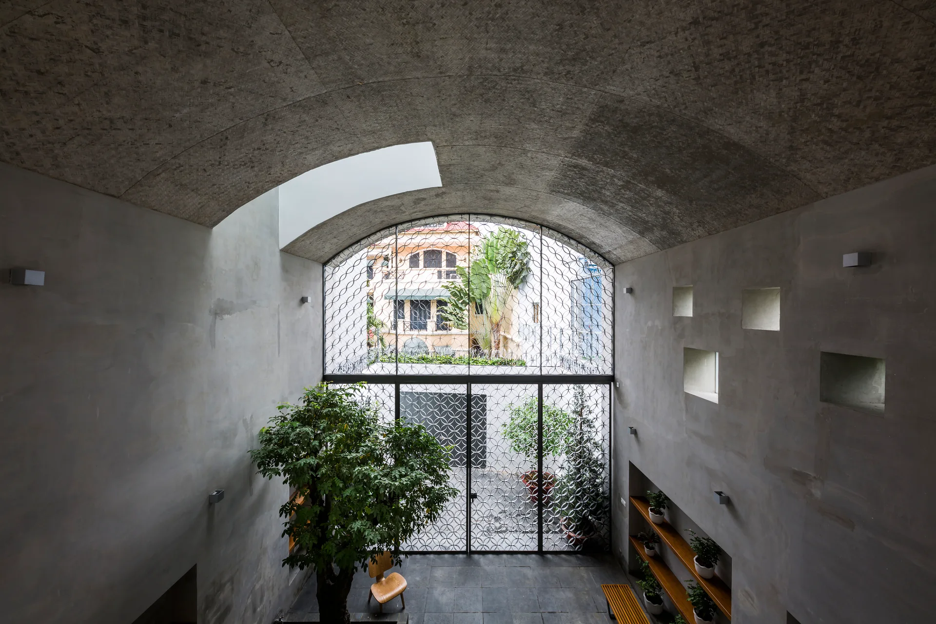 A minimalist concrete entry space with a vaulted ceiling, a large arched window with a geometric grille, and a potted tree.