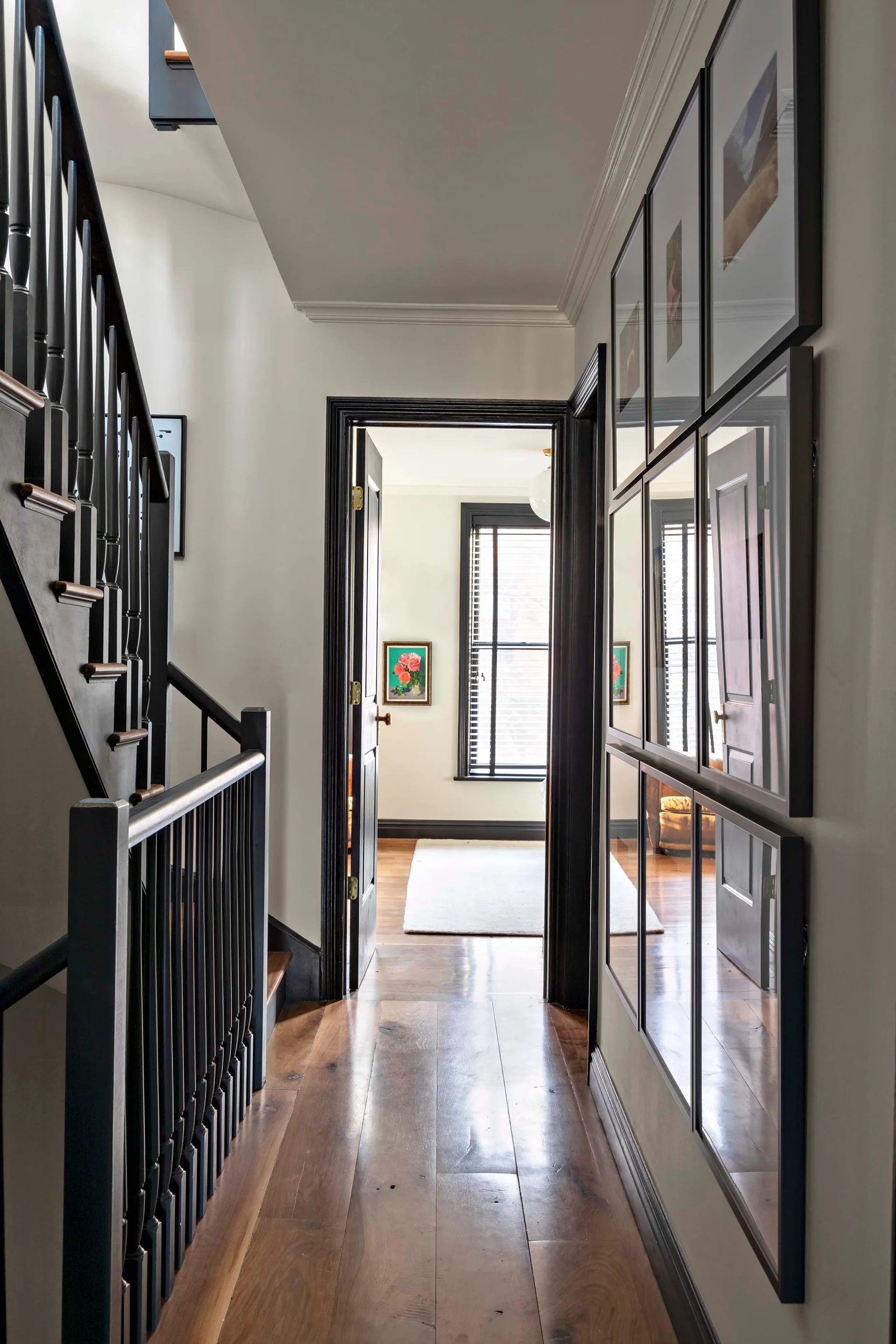A narrow hallway features white walls, dark wood floors, black door trim, and a black staircase railing opposite a grid of framed pictures.