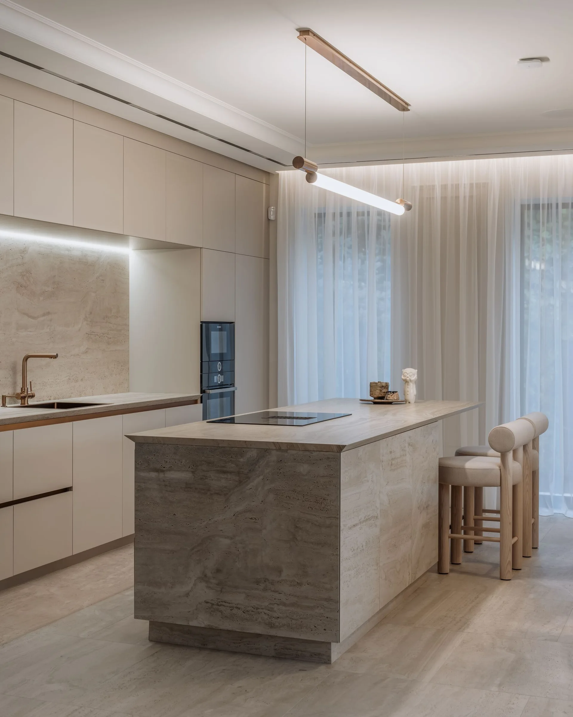 A minimalist cream kitchen features a travertine stone island with two upholstered stools, matching flat-panel cabinetry, and sheer window curtains.