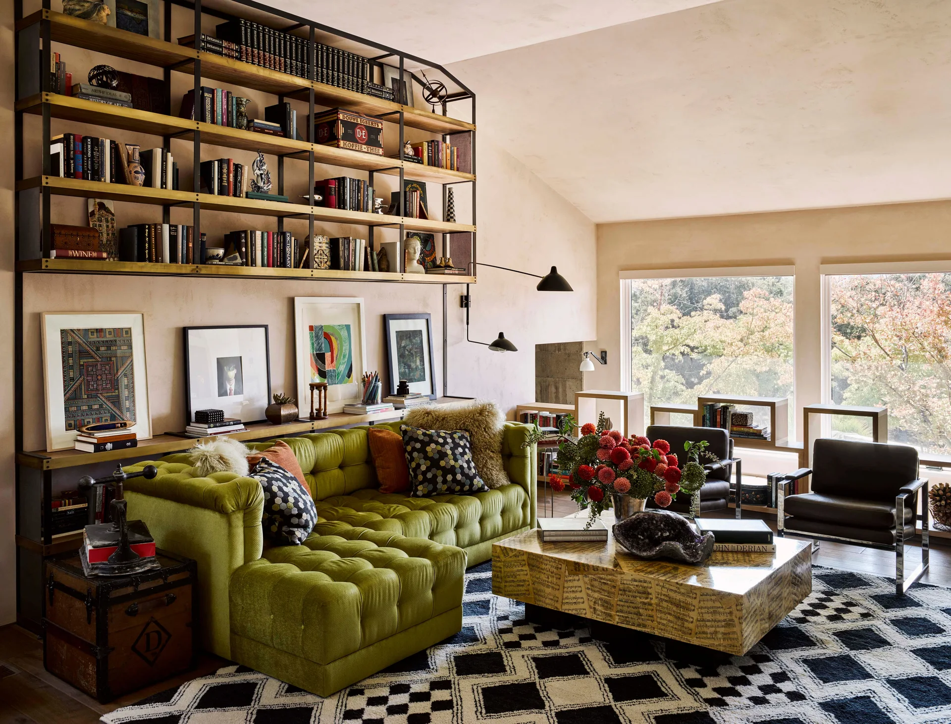 An eclectic living room features a tufted olive green sectional sofa, a black and white geometric rug, and a large metal wall shelving unit.
