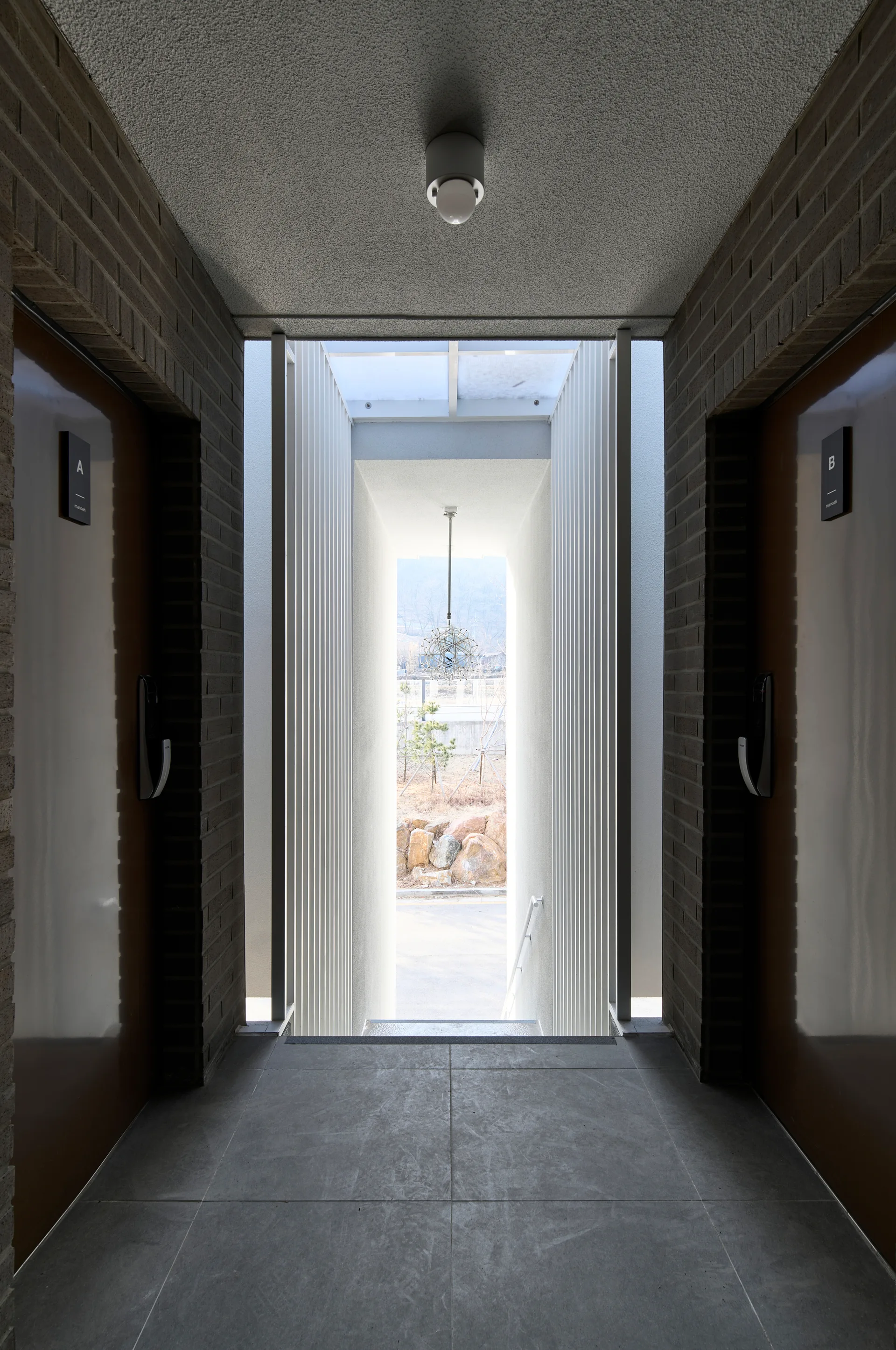 A symmetrical hallway with brown doors set in brick walls leads to a bright passage lined with vertical white slats and a hanging pendant light.