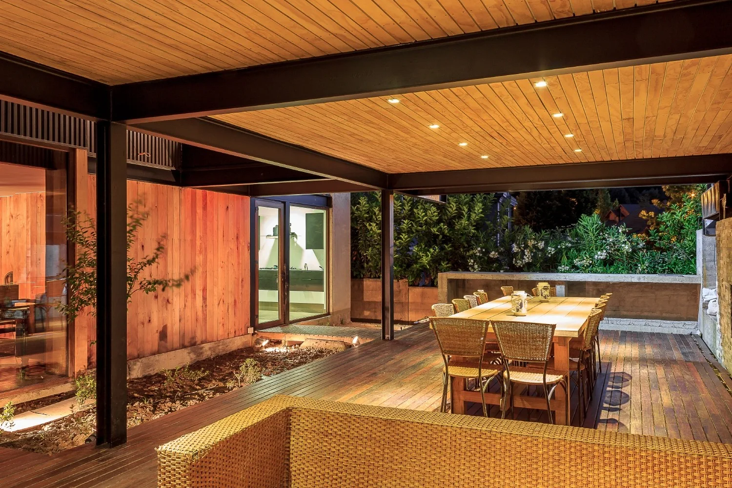 A covered wooden deck at night featuring a large dining table with woven chairs under a lit paneled ceiling with black steel beams.